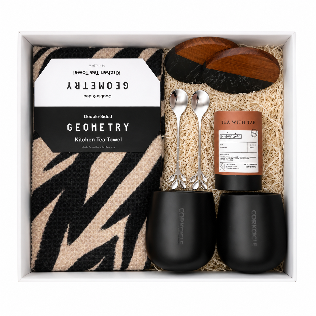 A gift box containing a kitchen tea towel with a black and beige zebra stripe pattern, two black cups labeled 'Corkcicle', two metal spoons with leaf-shaped handles, a cylindrical container of tea labeled 'Tea With Tae', and two black and wood coasters, all arranged on a bed of natural-colored shredded paper.