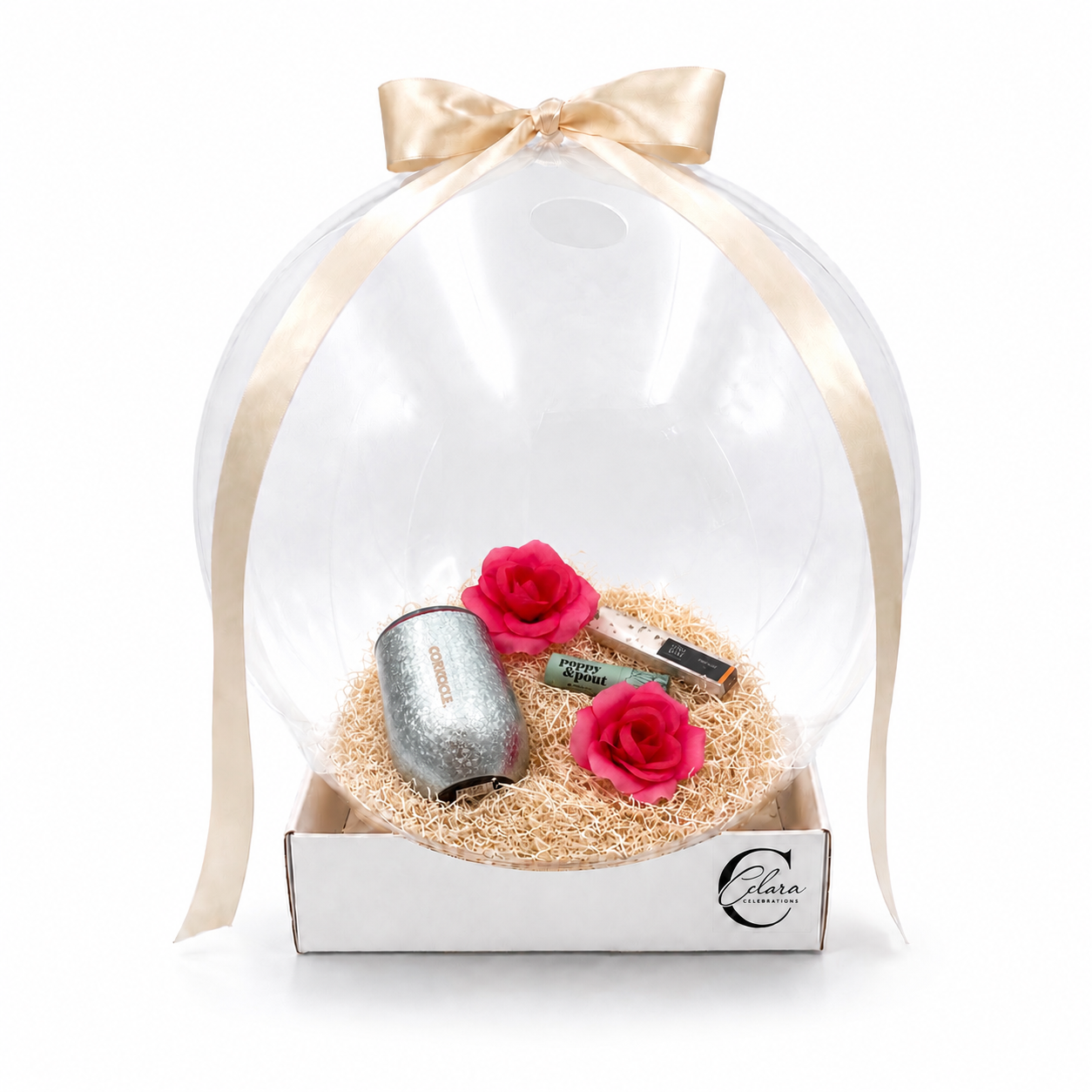 Clear gift box with a gold ribbon, containing pink roses, a silver candle, and small boxes of chocolates, resting on a bed of natural filler inside a white box labeled 'Clara Celebrations.'
