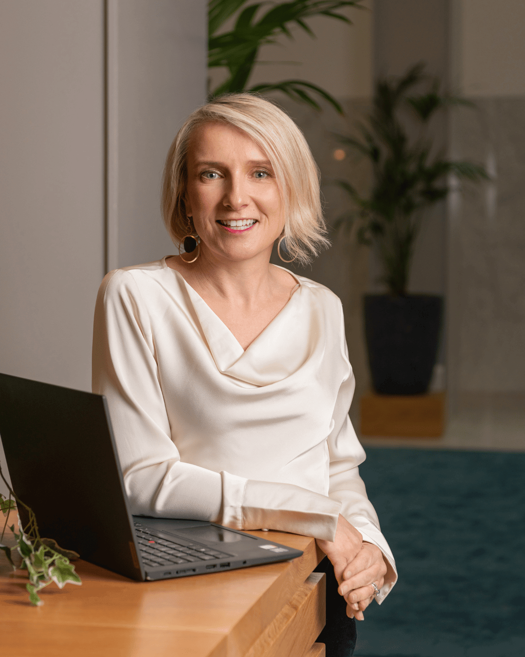 A woman with short blonde hair, wearing a white satin blouse with a draped neckline, smiling and standing behind a wooden desk with a black laptop open. There are indoor plants and a gray wall in the background.