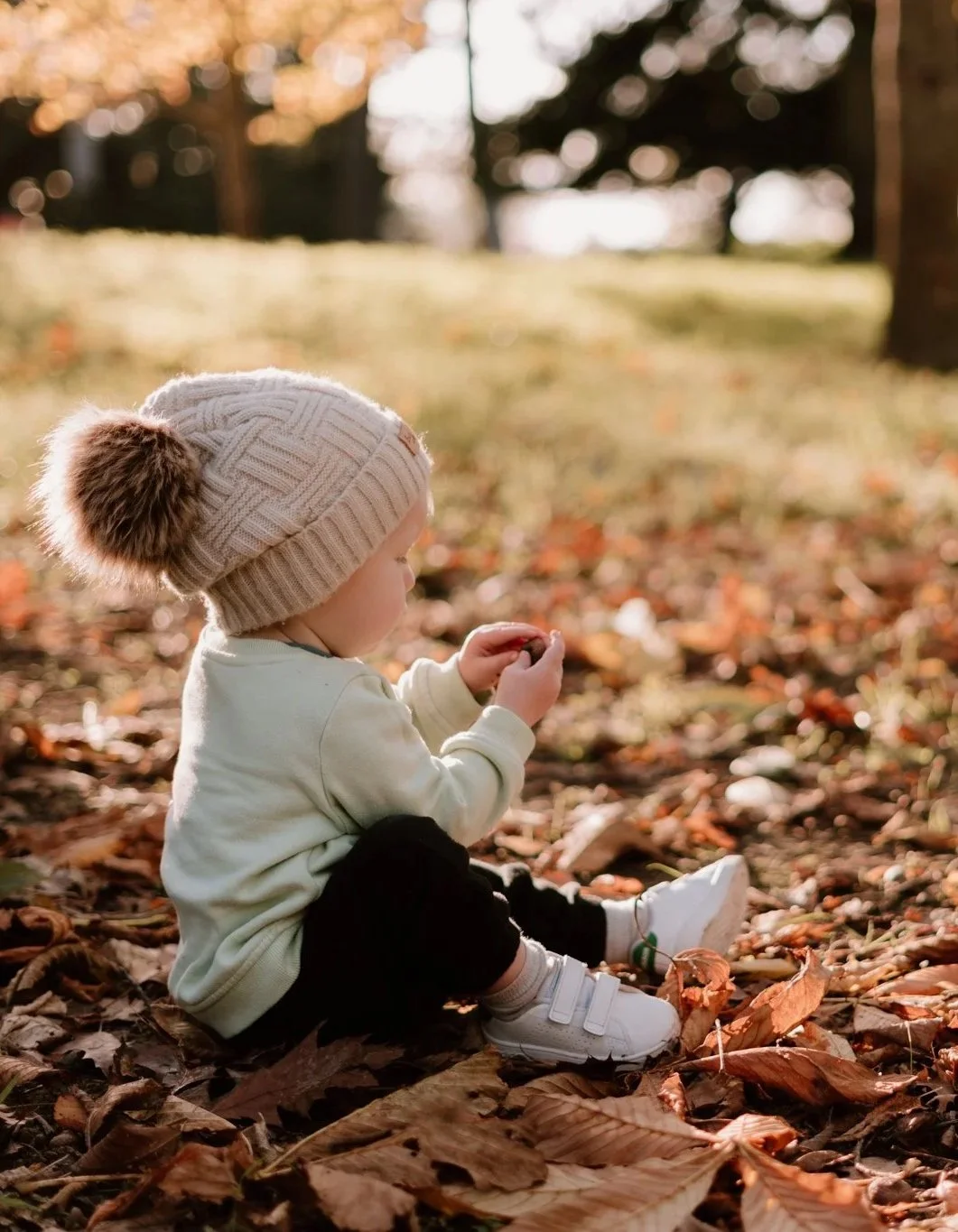A young child sitting on the ground covered with fallen autumn leaves, wearing a knit hat with a pom-pom, a cream-colored jacket, black pants, and white shoes, holding a leaf in their hands, outdoors in a park or wooded area during fall.