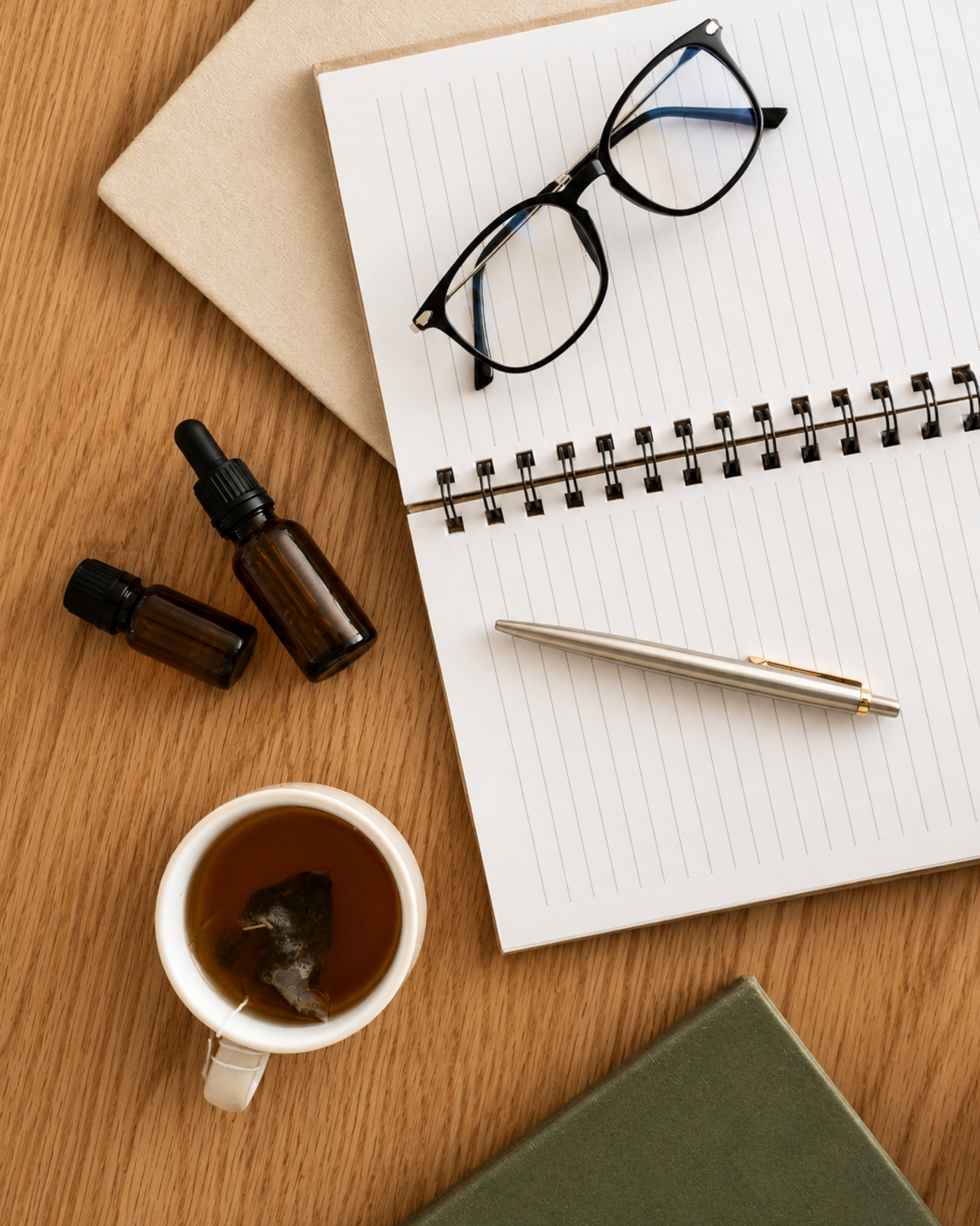 A wooden desk with a pair of black eyeglasses on a spiral notebook, a pen, a cup of tea, two small brown bottles, a beige tote bag, and a closed green book.