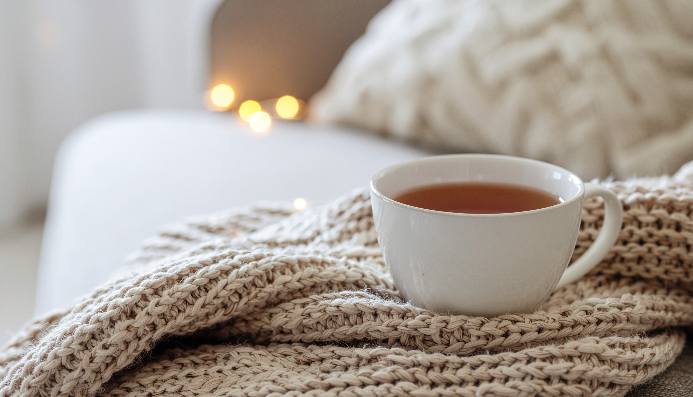 A white ceramic mug filled with tea rests on a chunky beige knit blanket, with a cozy, softly lit background featuring blurred warm lights and a textured pillow.