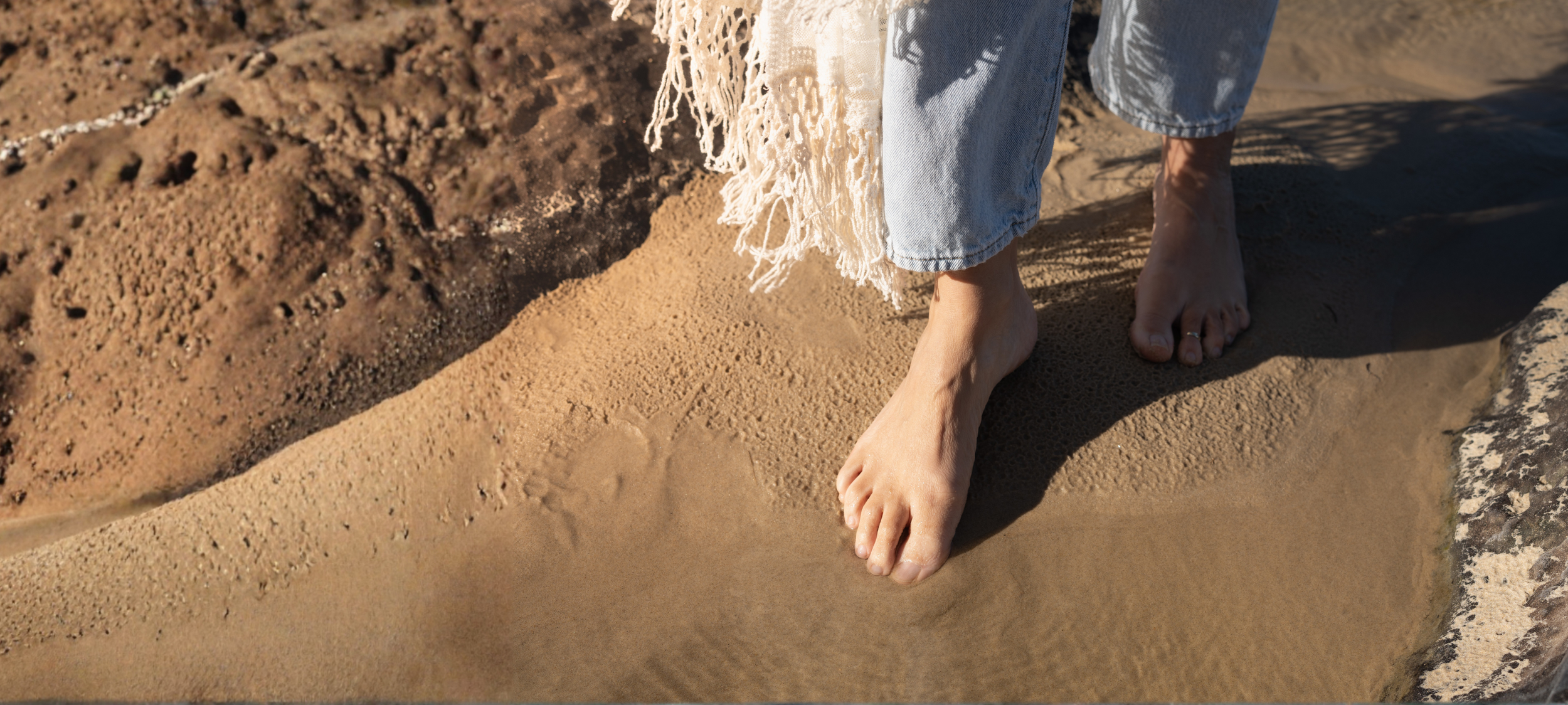 Person barefoot on sandy beach, wearing ripped jeans and a fringed white shawl.
