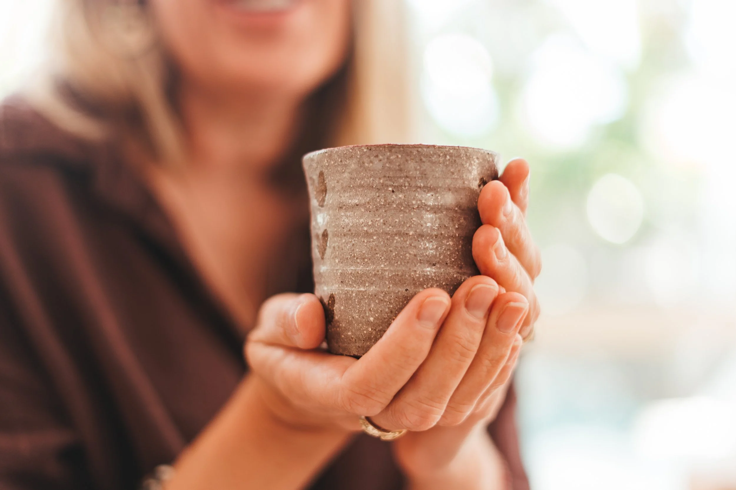 A woman holding a textured ceramic cup with both hands, near her chest, blurred background with natural light.