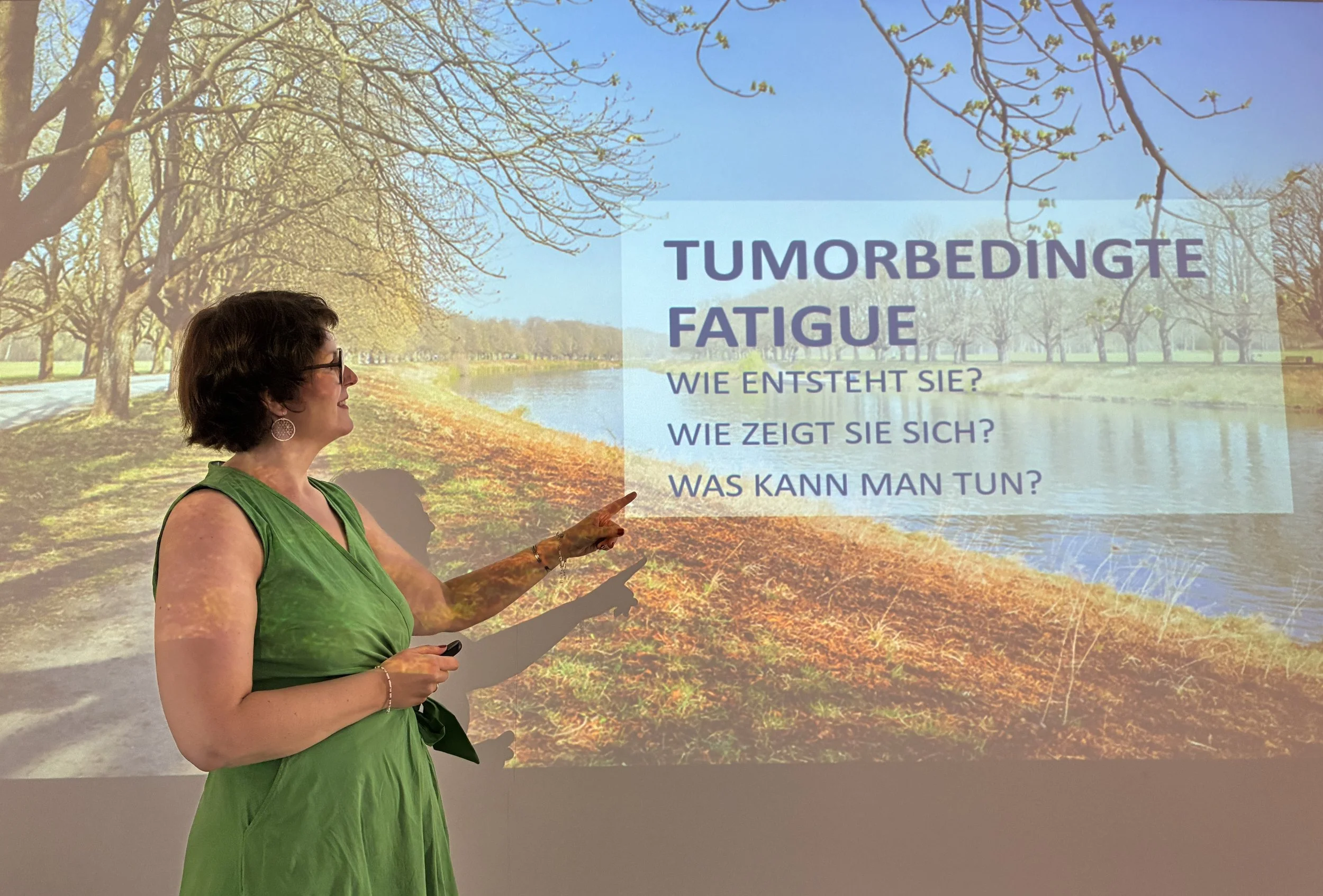 Eine Frau in grüner Kleidung zeigt auf eine Präsentation mit dem Titel 'Tumorbedingt Fatigue' vor einem Hintergrund eines Parks mit Bäumen und einem Wasserarm des Parks.