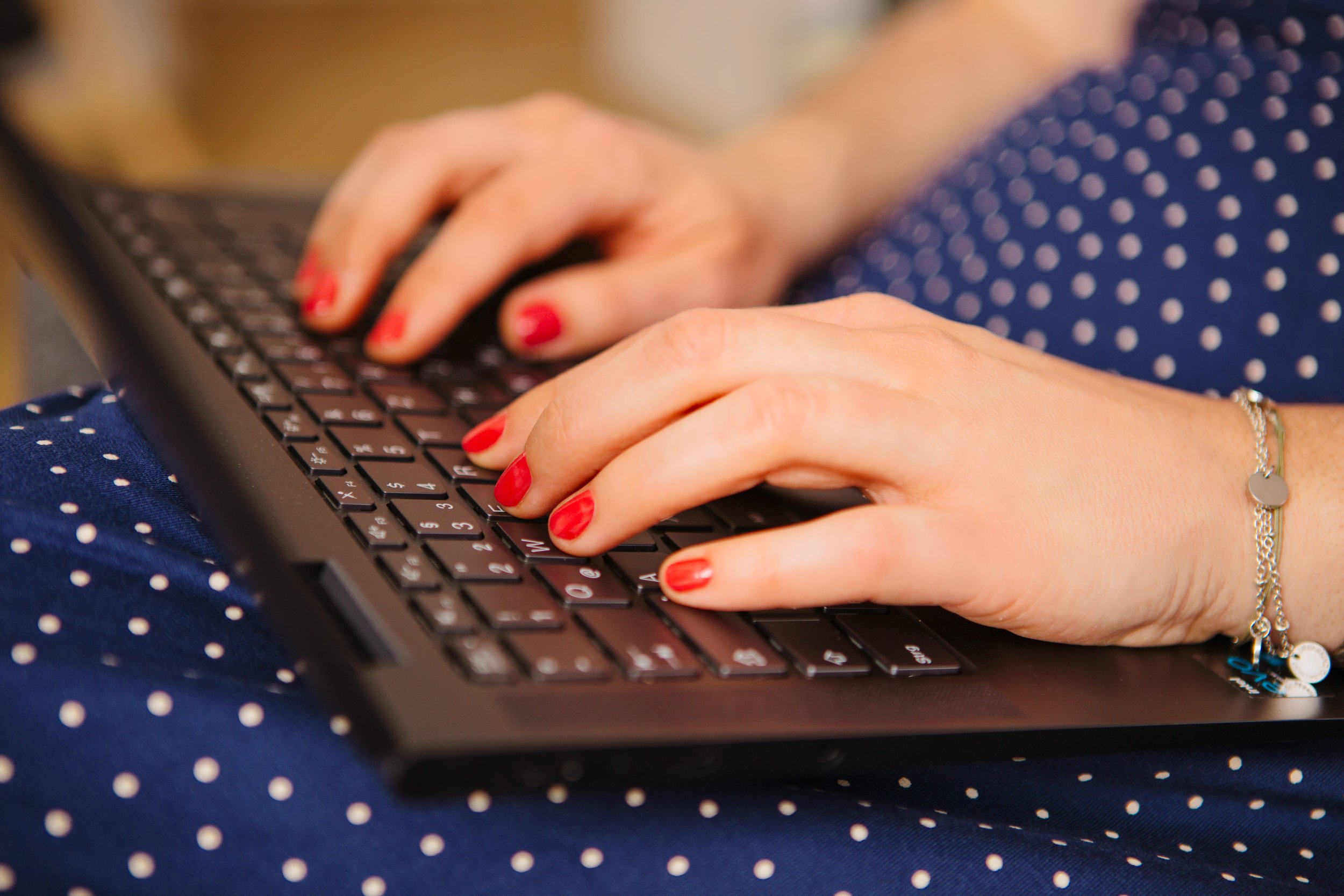 Nahaufnahme von Händen beim Tippen auf einer schwarzen Tastatur, getragen von einer Person mit rotem Nagellack, blauem Kleid mit weißen Punkten und Armband.