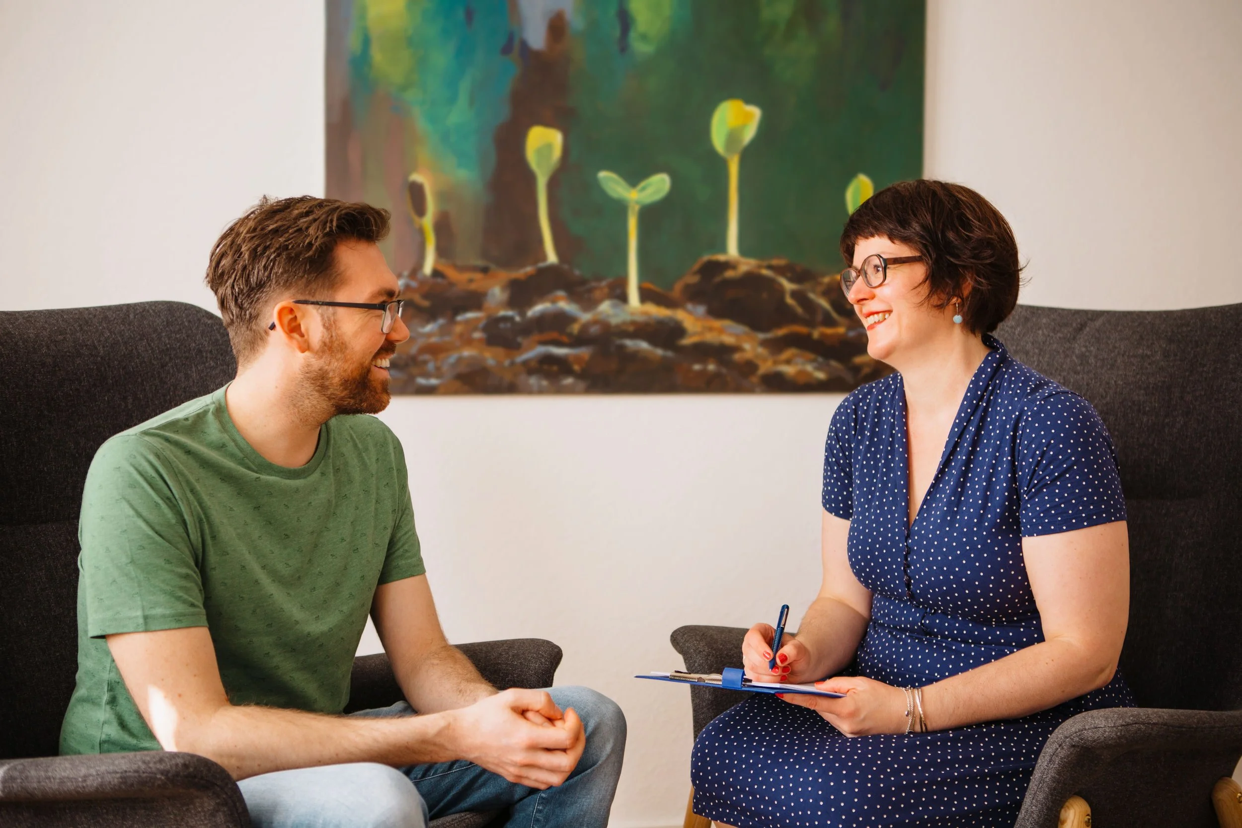 Zwei Menschen, ein Mann mit Brille und grüner T-Shirt, und eine Frau mit Brille und blauer Kleid mit weißen Punkten, sitzen sich gegenüber auf einem Sofa und unterhalten sich lächelnd. Im Hintergrund ist ein Gemälde mit jungen Pflanzen zu sehen.