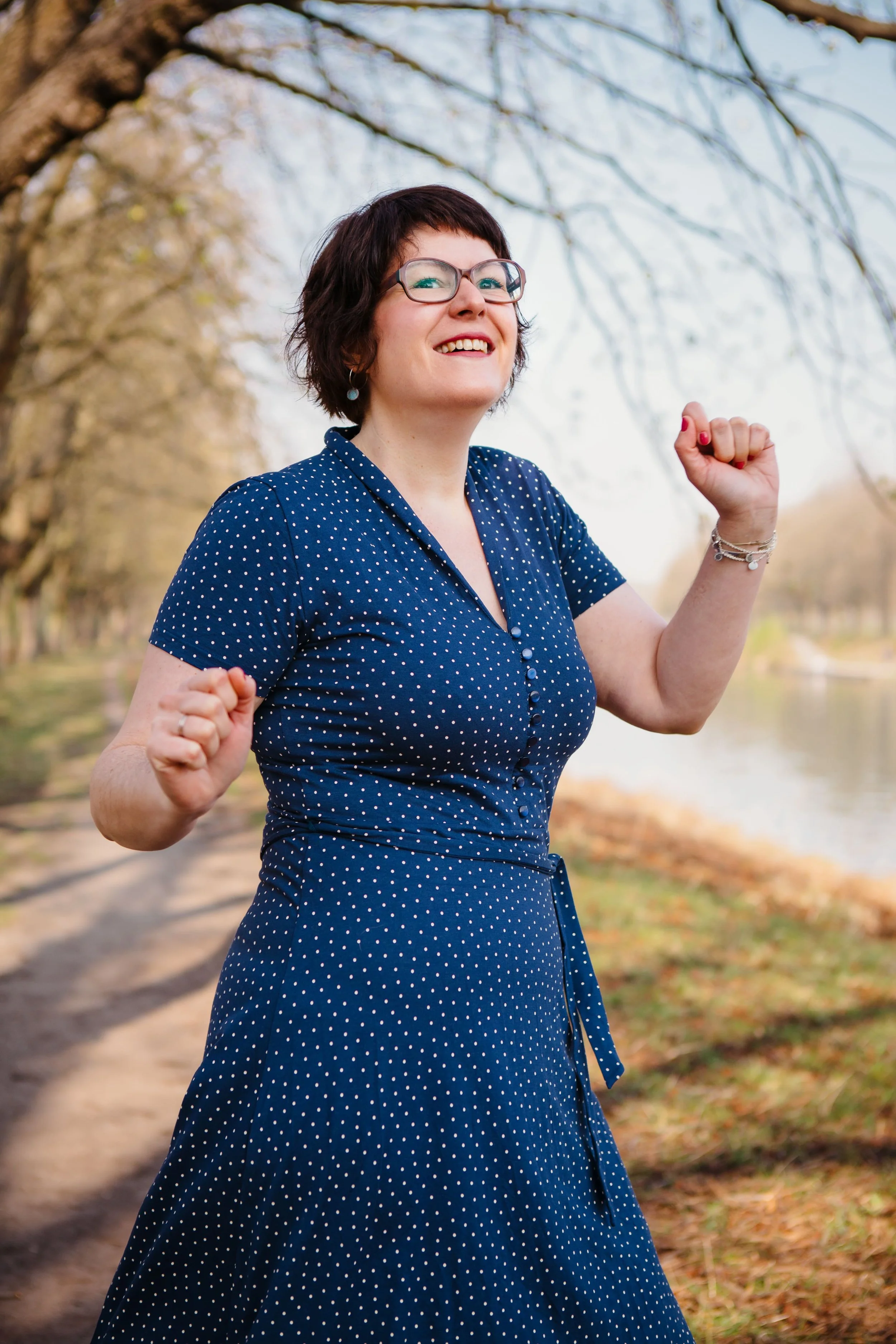 Eine lächelnde Frau mit Brille, die in einem blauen Kleid mit weißen Punkten im Freien an einem sonnigen Tag steht und die Arme hoch macht.
