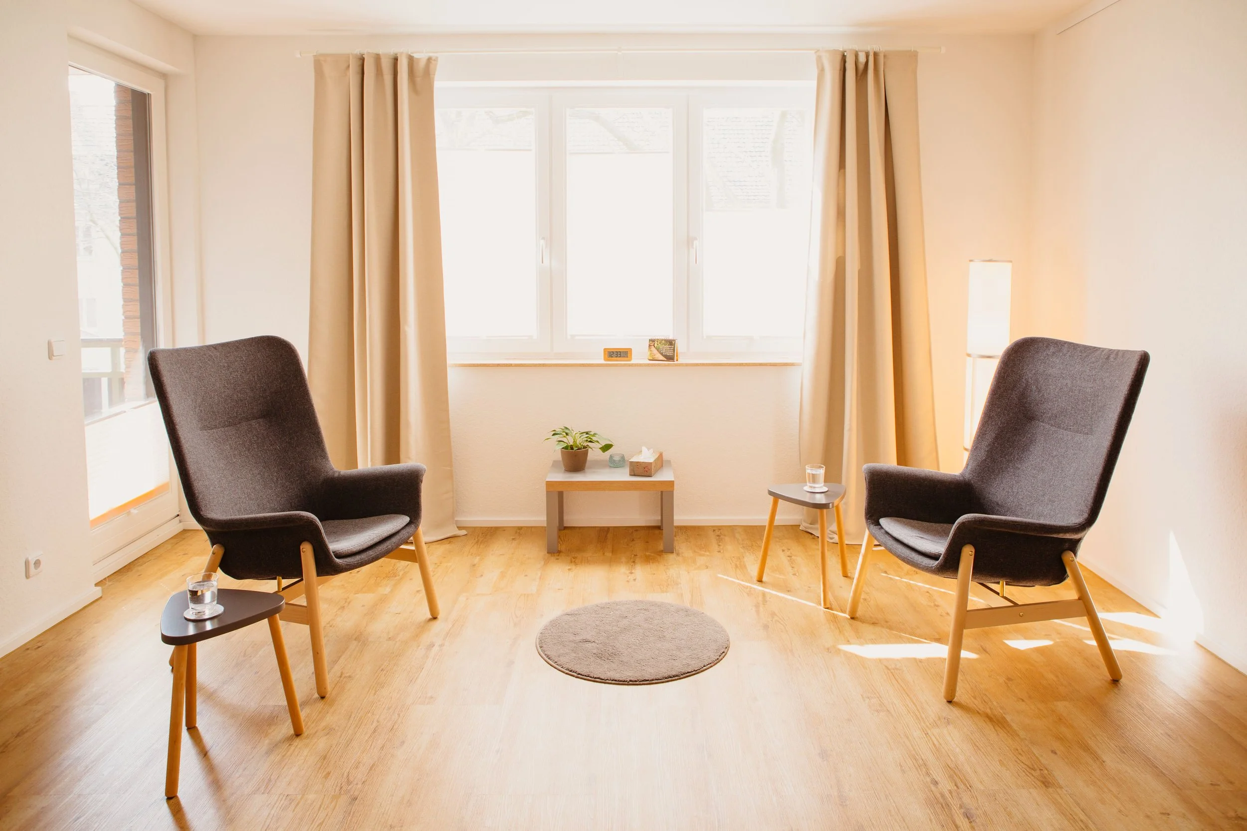 Ein Wohnzimmer mit zwei schwarzen Sesseln, eine kleine Tabelle mit Pflanzen und einer Tasse, große Fenster mit weißen Vorhängen, Holzboden, Tageslicht