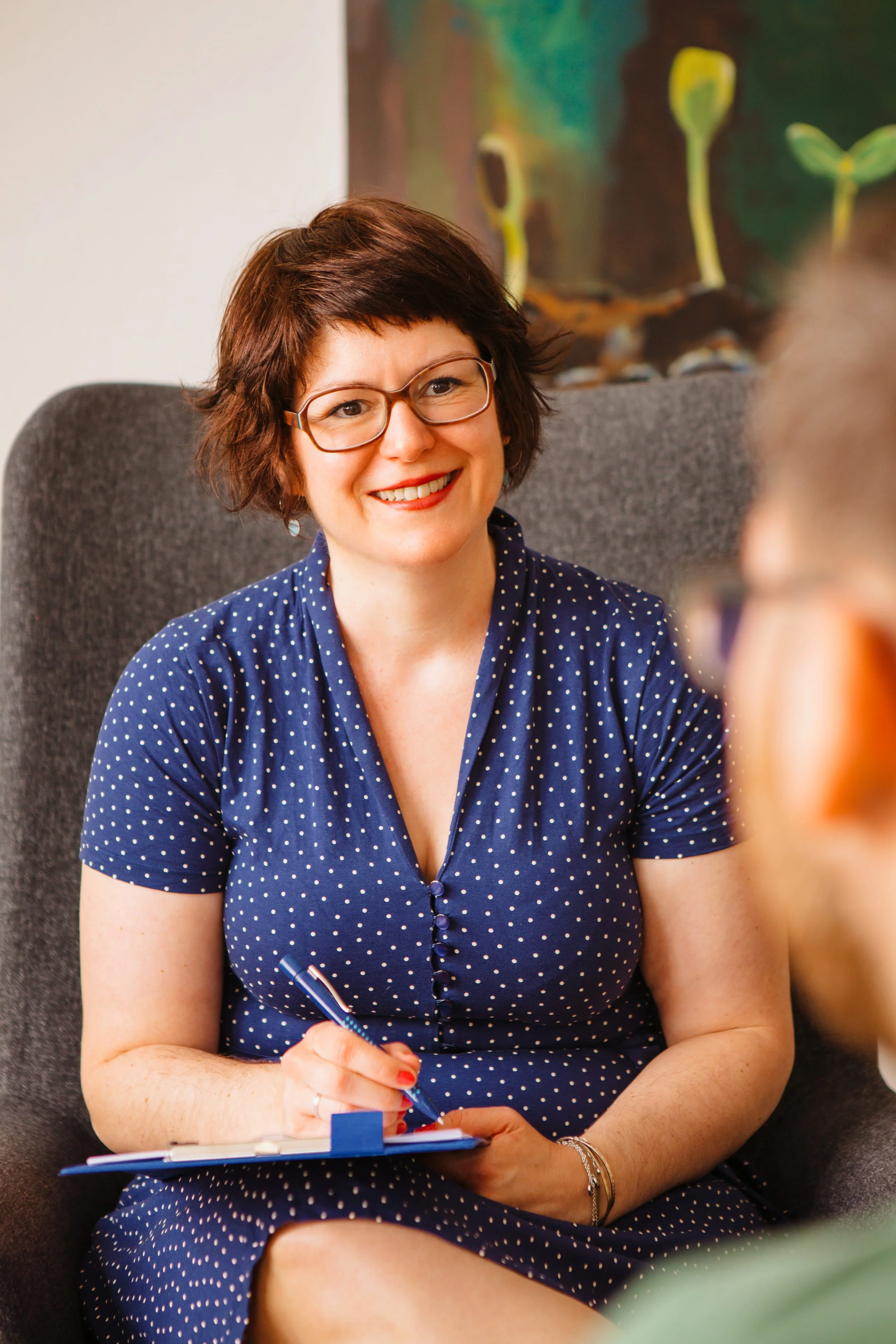 Eine lachende Frau mit Brille, die rot geschminkt ist, sitzt auf einem Stuhl mit grauem Bezug. Sie trägt ein dunkelblaues Kleid mit weißen Punkten und hält einen blauen Stift in der Hand, während sie Notizen auf einem Notizblock macht.