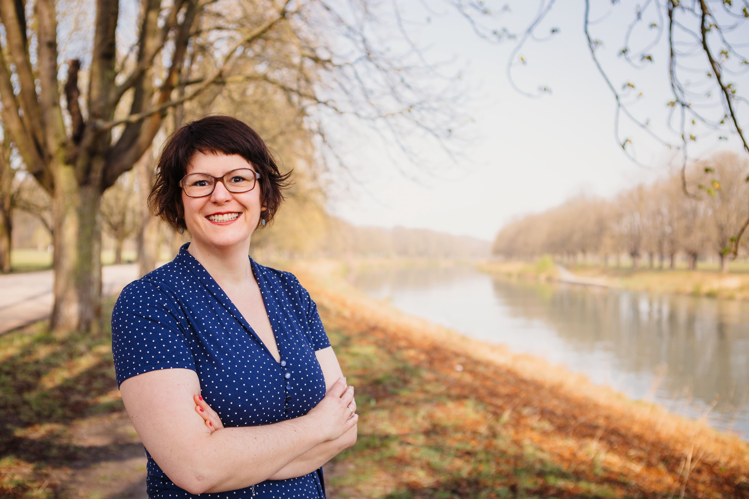 Eine Frau mit kurz braunen Haaren, Brille und einem blauen Kleid mit weißen Punkten steht in einem Park an einem Fluss, umgeben von Bäumen, die im Herbst ihre Blätter verlieren.