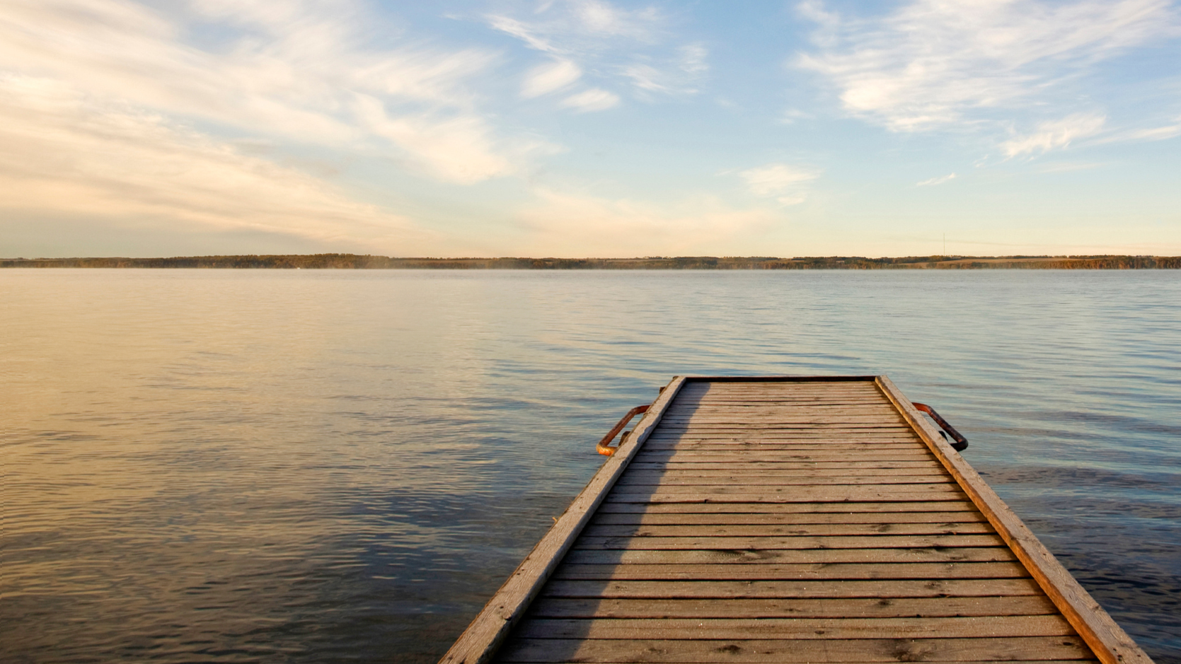 Holzsteg führt zu einem ruhigen See bei Sonnenuntergang, Himmel mit Wolken am Horizont.