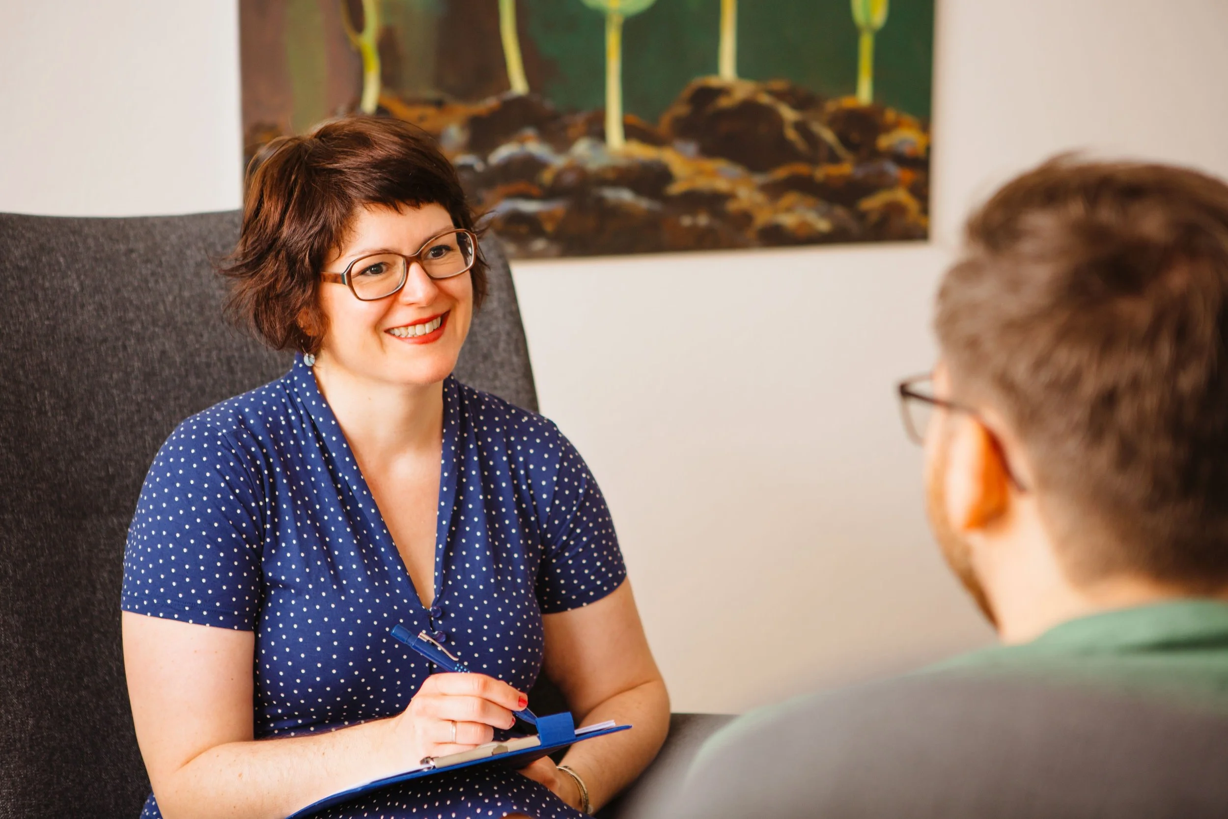 Eine Frau mit Brille lächelt während eines Gesprächs mit einem Mann, der nur teilweise im Bild zu sehen ist, in einem Büro. Im Hintergrund ist ein abstraktes Bild an der Wand zu sehen.