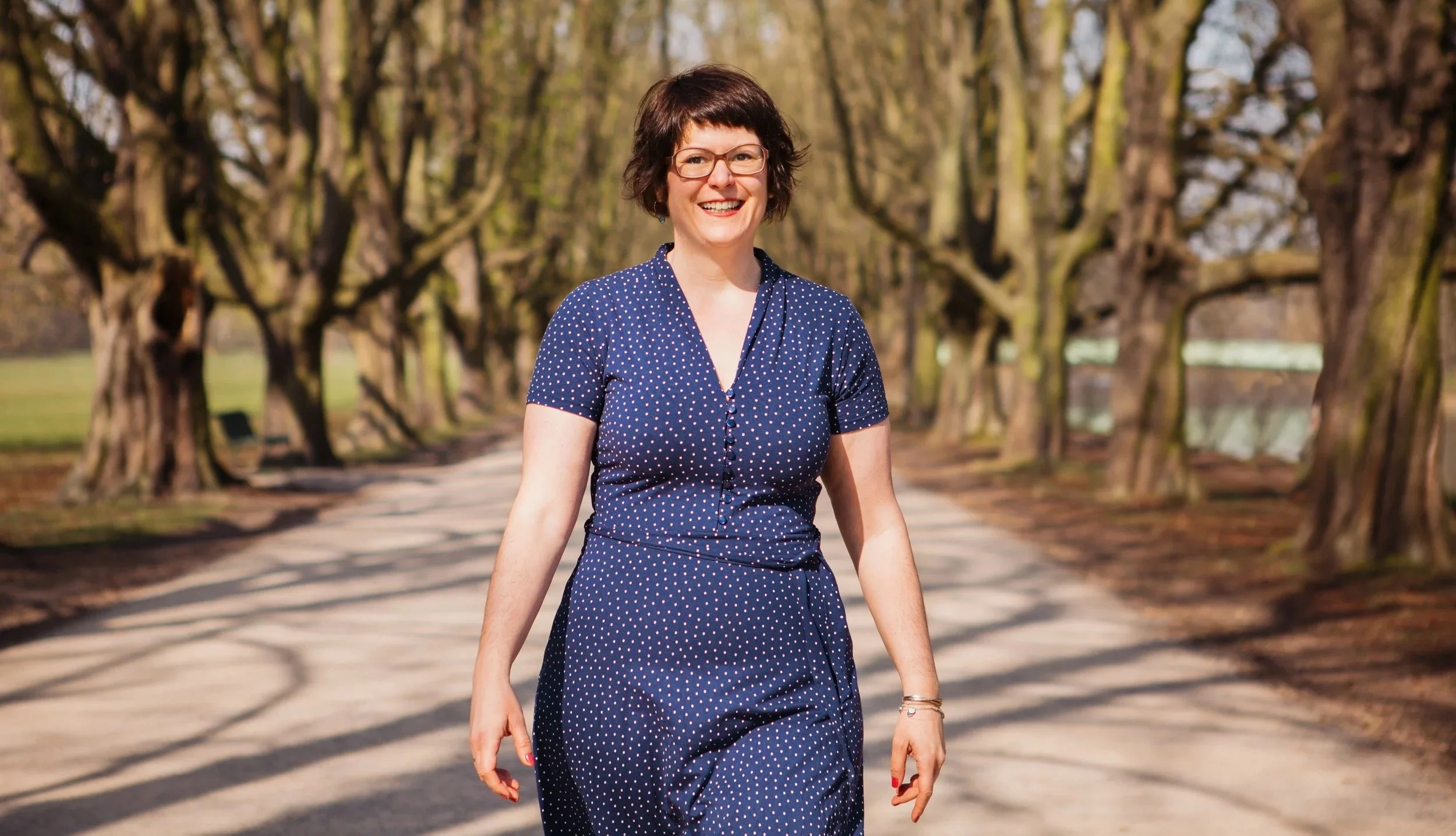 Eine lachende Frau mit Brille und dunkelbraunem, relativ kurzem Haar läuft auf einer Baumallee im Freien.