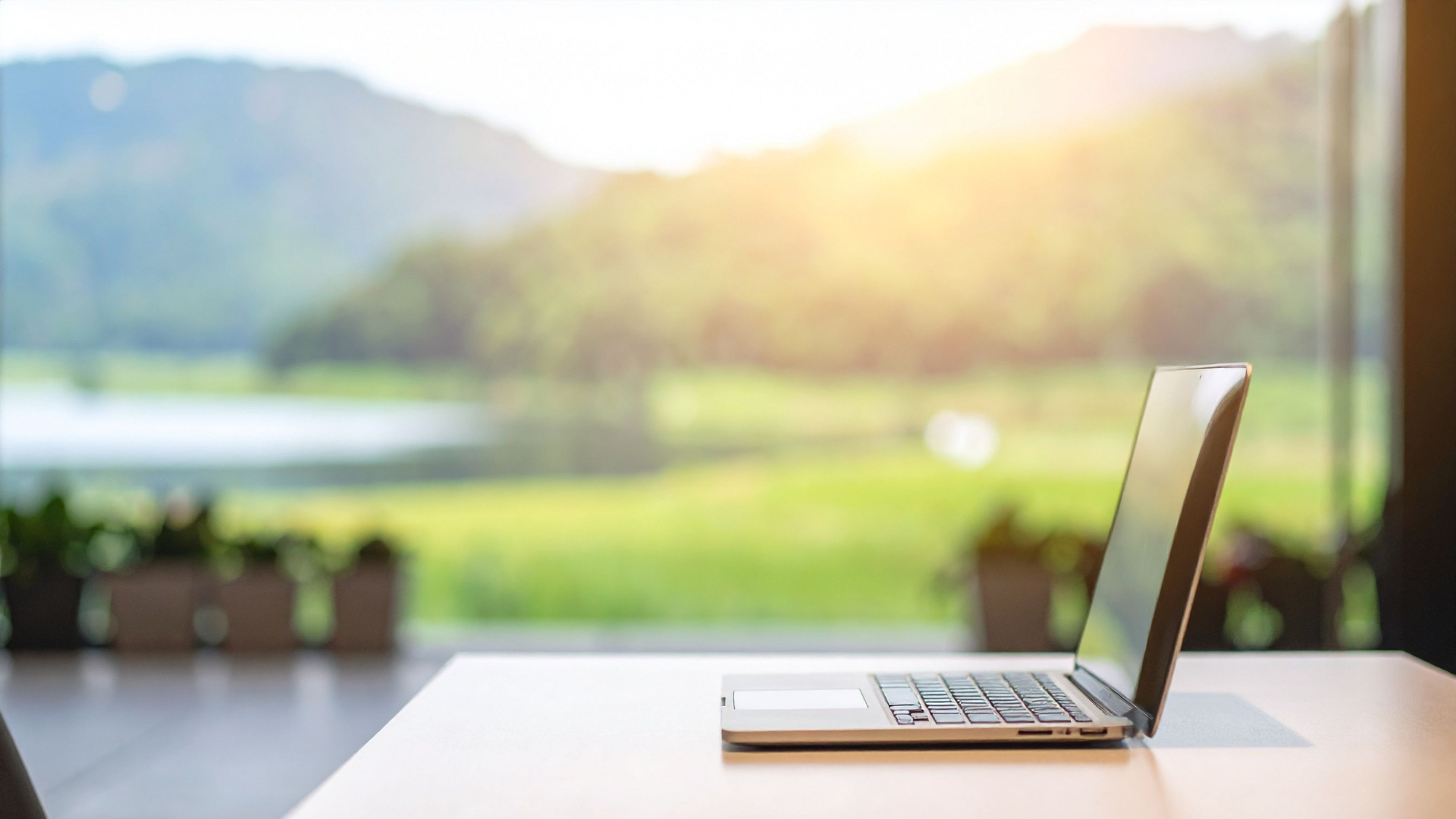 Ein aufgeschlagener Laptop auf einem weißen Tisch vor einem großen Fenster, durch das eine grüne Landschaft mit Bergen bei Sonnenuntergang sichtbar ist.