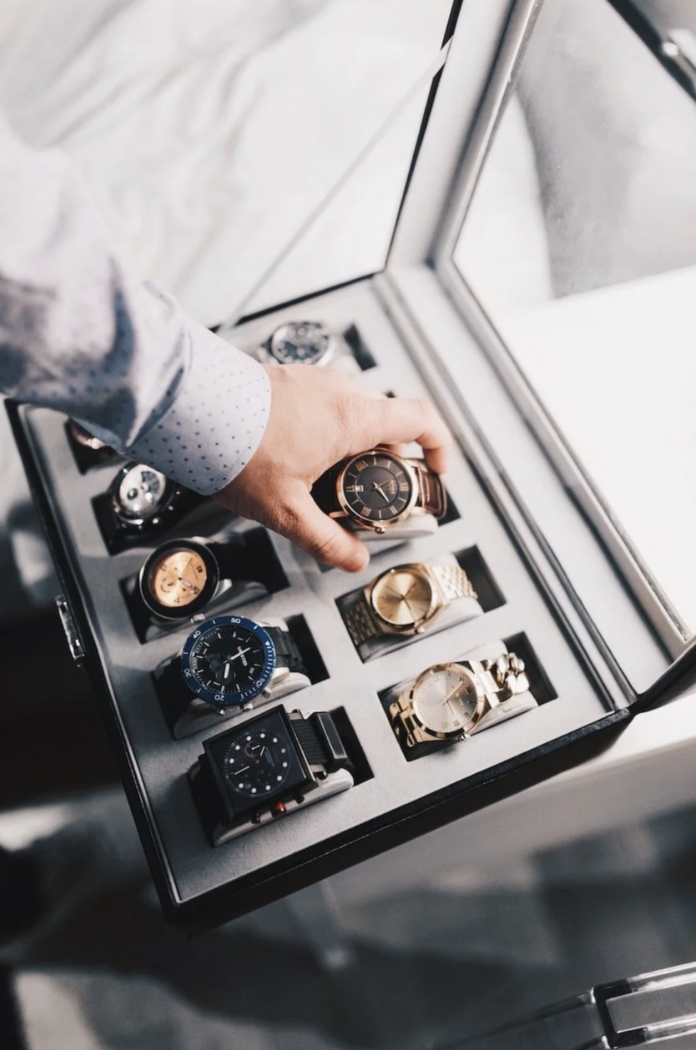 A person with a polka dot shirt is selecting a black and gold wristwatch from a watch display case containing various luxury watches.