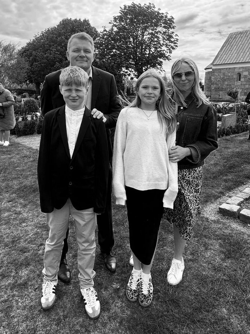 A group of four people, two adults and two children, standing outdoors on grass, smiling at the camera. The man and woman are standing behind the children. The woman is wearing sunglasses and a denim jacket, the man is in a suit, the boy is in a blazer and light pants, and the girl is in a sweater and patterned shoes. In the background, there are other people, trees, and a stone building.
