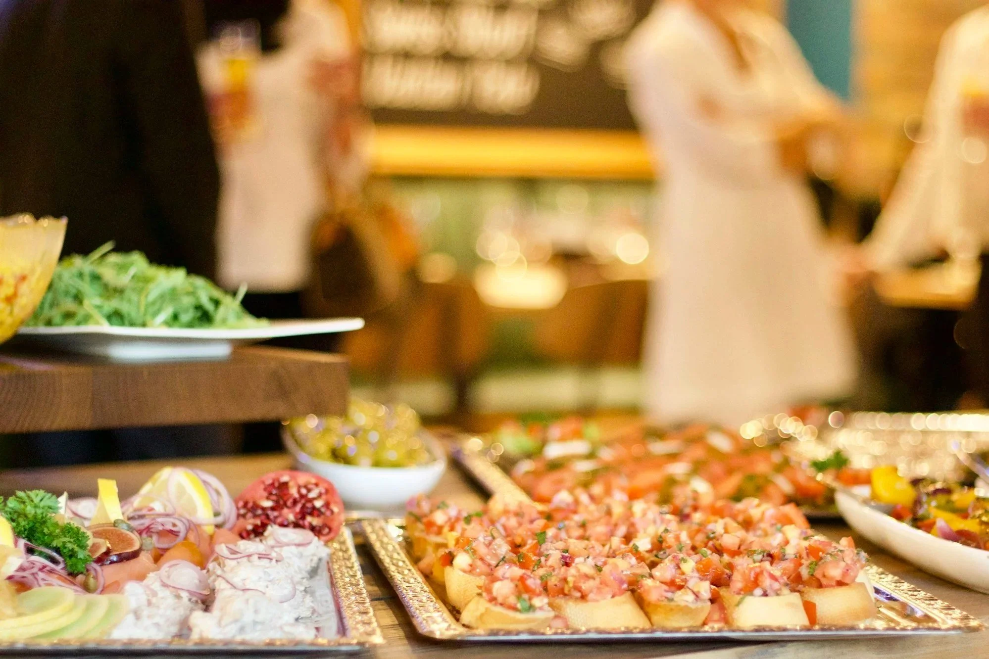 A buffet table with various appetizers including sliced meats, vegetables, and bread with toppings, displayed on gold trays and plates in a festive setting.