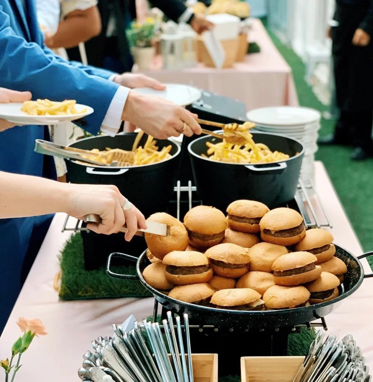 People serving pasta and sliders at a buffet table during an event.