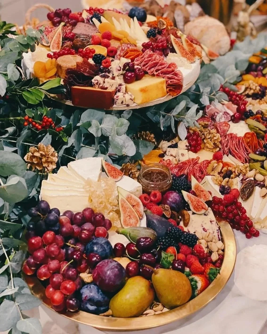 A large tray of assorted cheeses, fresh fruits, berries, nuts, and charcuterie on a decorated table, with greenery and additional food trays in the background.