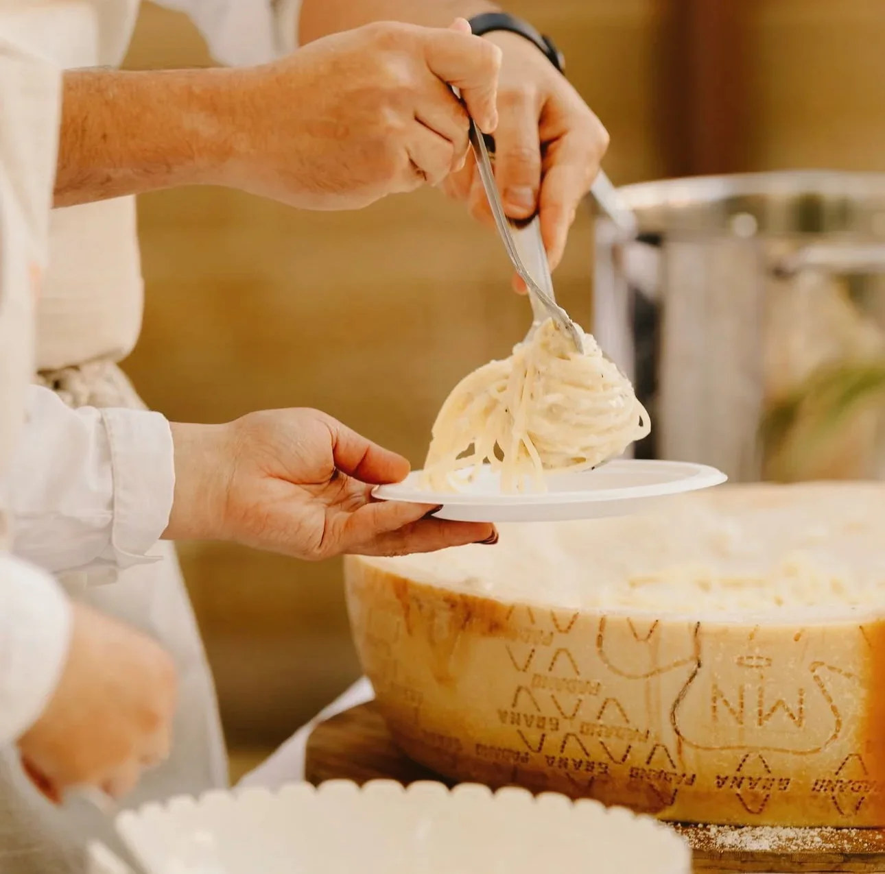 Person serving spaghetti pasta onto a plate from a large cheese wheel filled with pasta.
