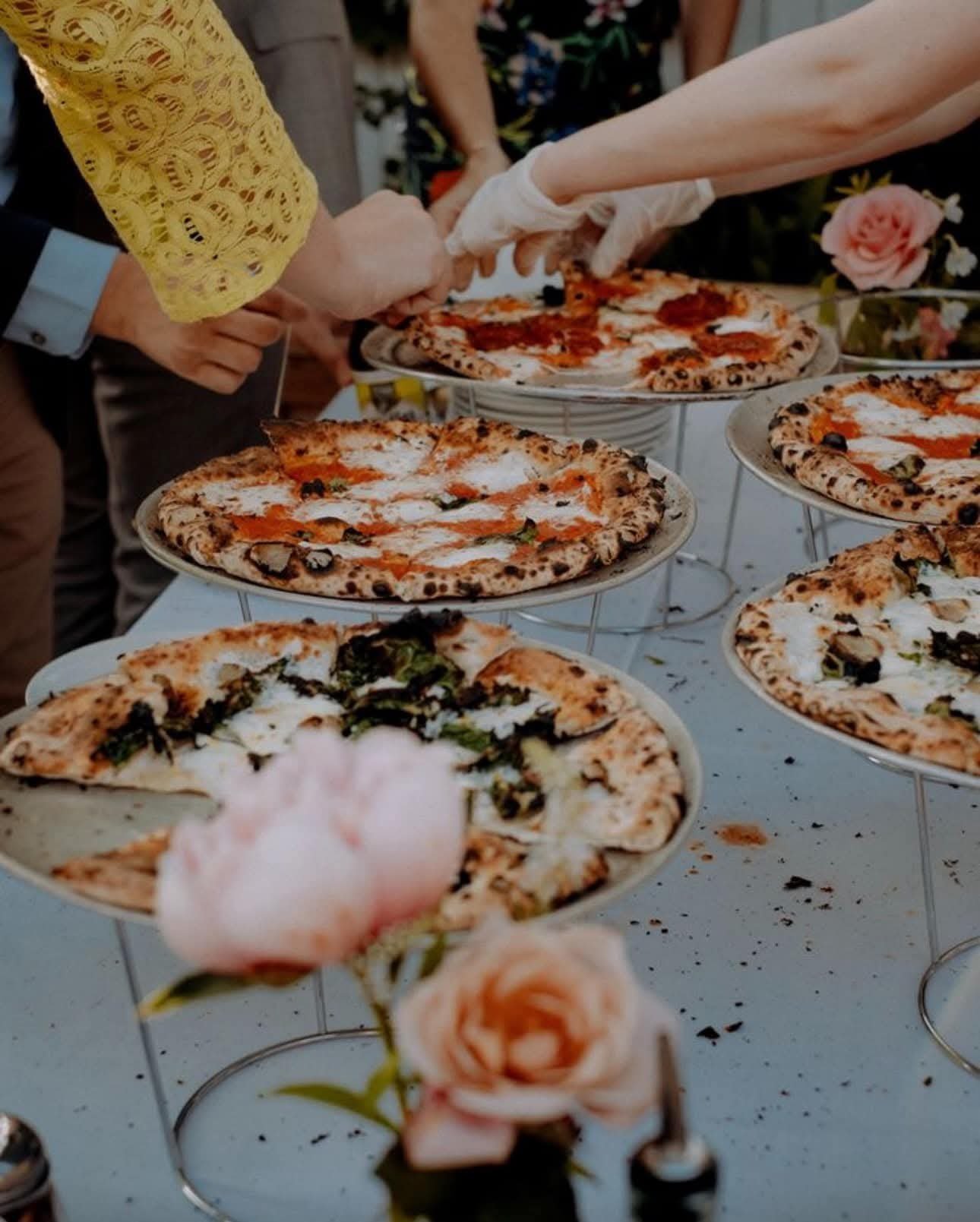 Multiple pizzas with various toppings on display at a gathering, with people serving and eating in the background.