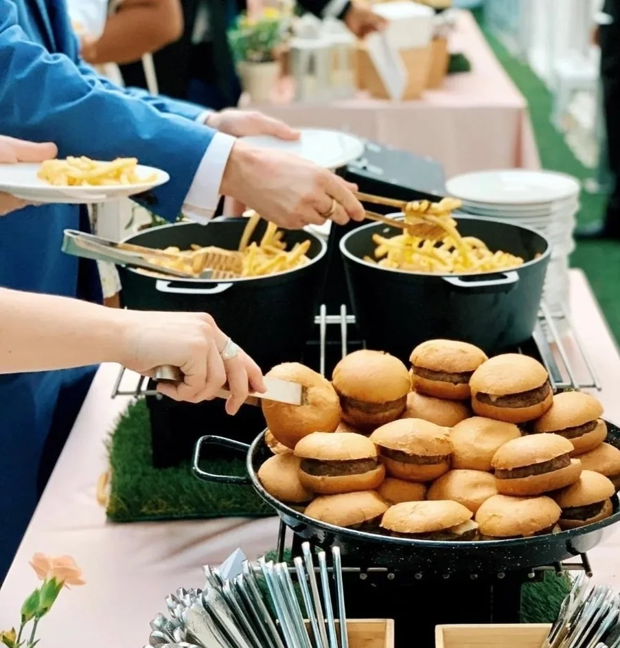 Buffet table with burgers, pasta, and plates at a gathering.