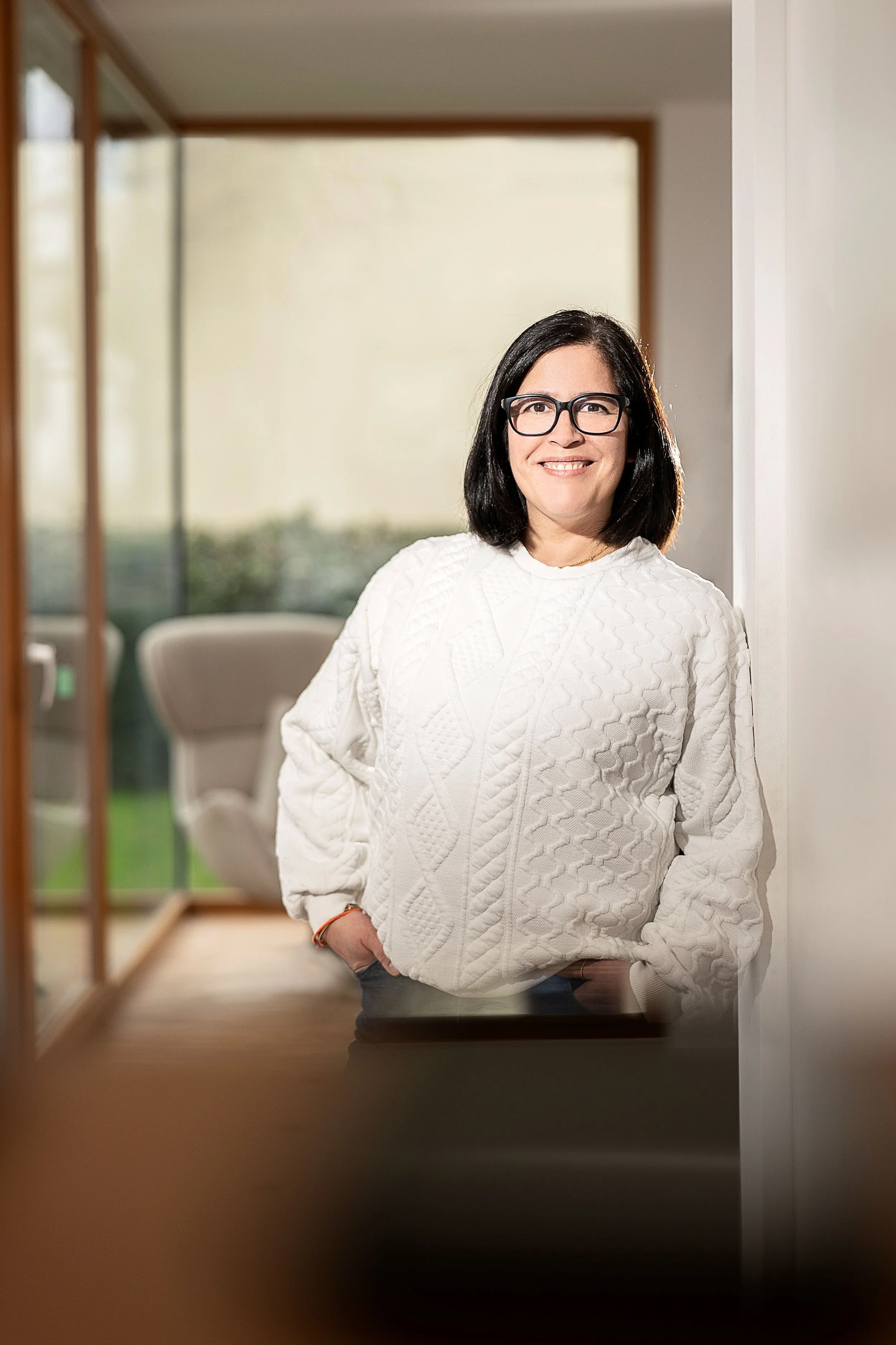 Frau mit schwarzen Haaren und Brille, trägt weiße Strickpullover, steht im Raum und lächelt.
