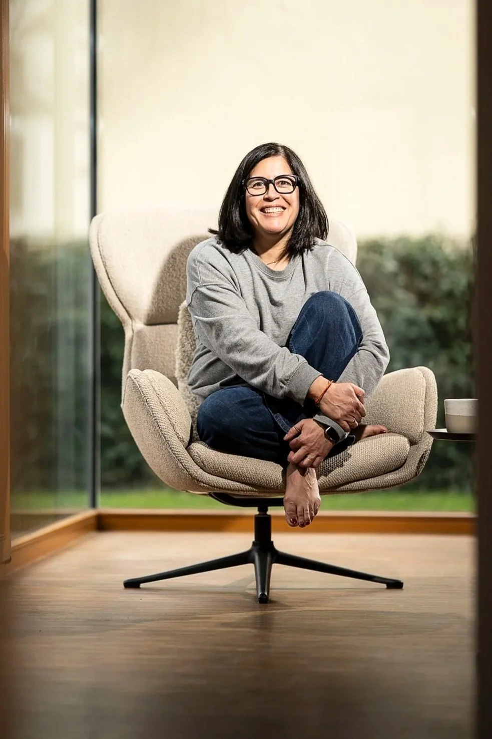 Frau mit dunklen Haaren, Brille und grauem Pullover sitzt auf einem modernen bequemen Stuhl vor einem Fenster, im Hintergrund grüner Garten, lächelnd, entspannt.