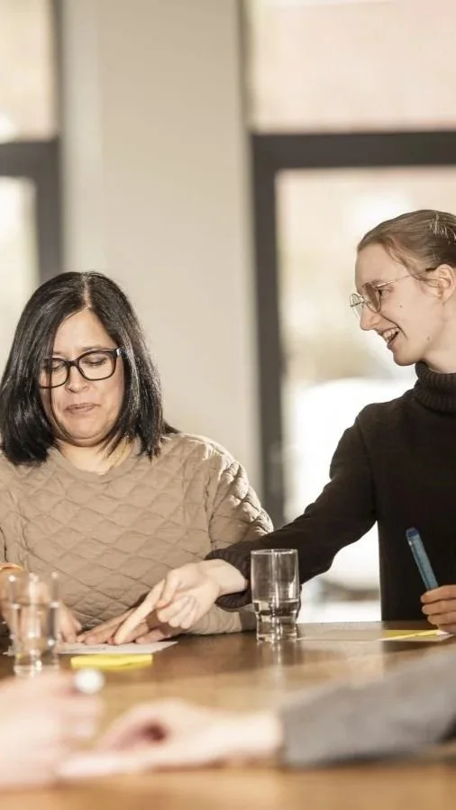 Zwei Frauen sitzen an einem Tisch im Gespräch, eine Frau legt ihre Hand auf die Hand der anderen, beide tragen Brillen.