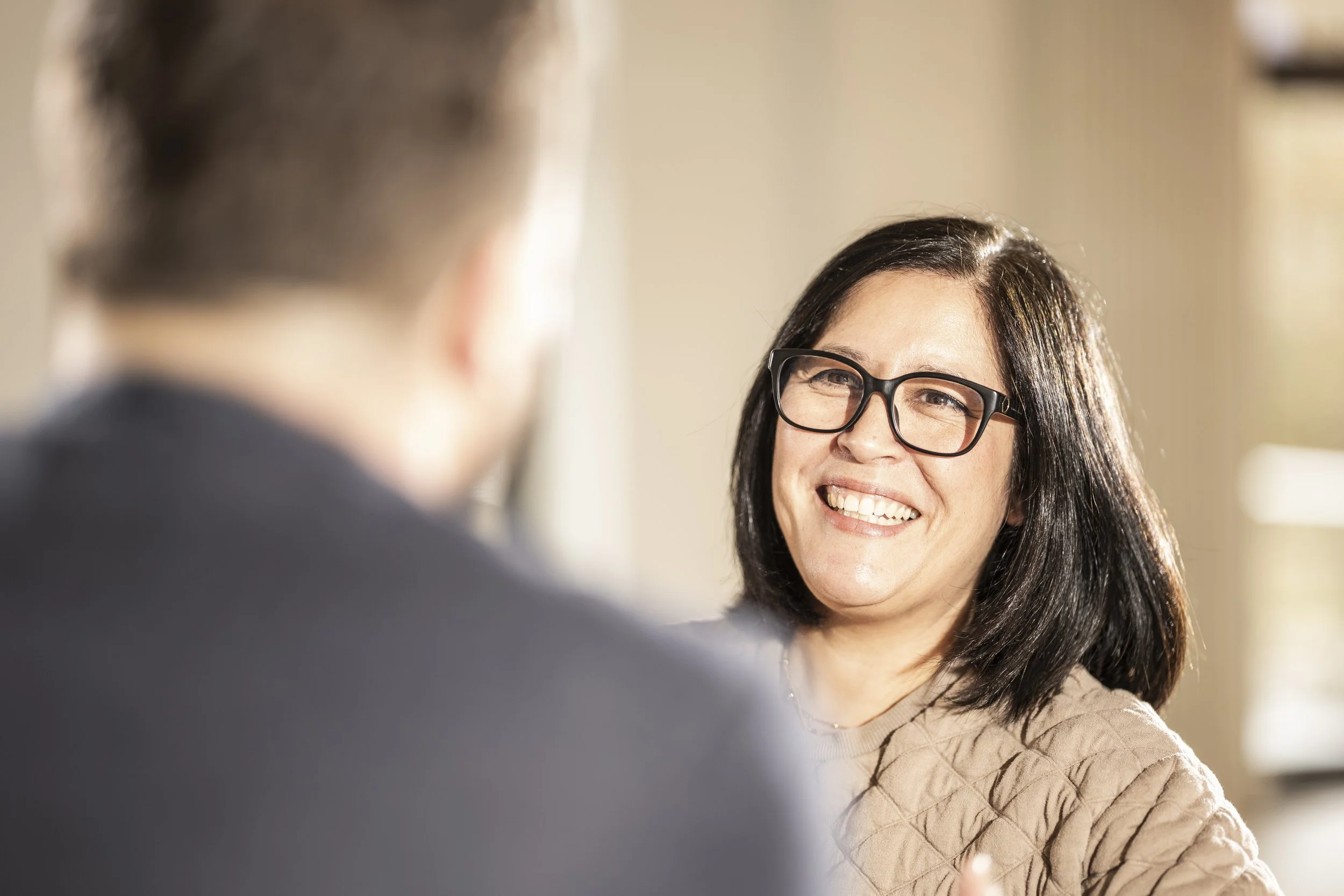 Frau mit dunklen Haaren und Brille lächelt während eines Gesprächs mit Mann, der im Vordergrund unscharf ist.