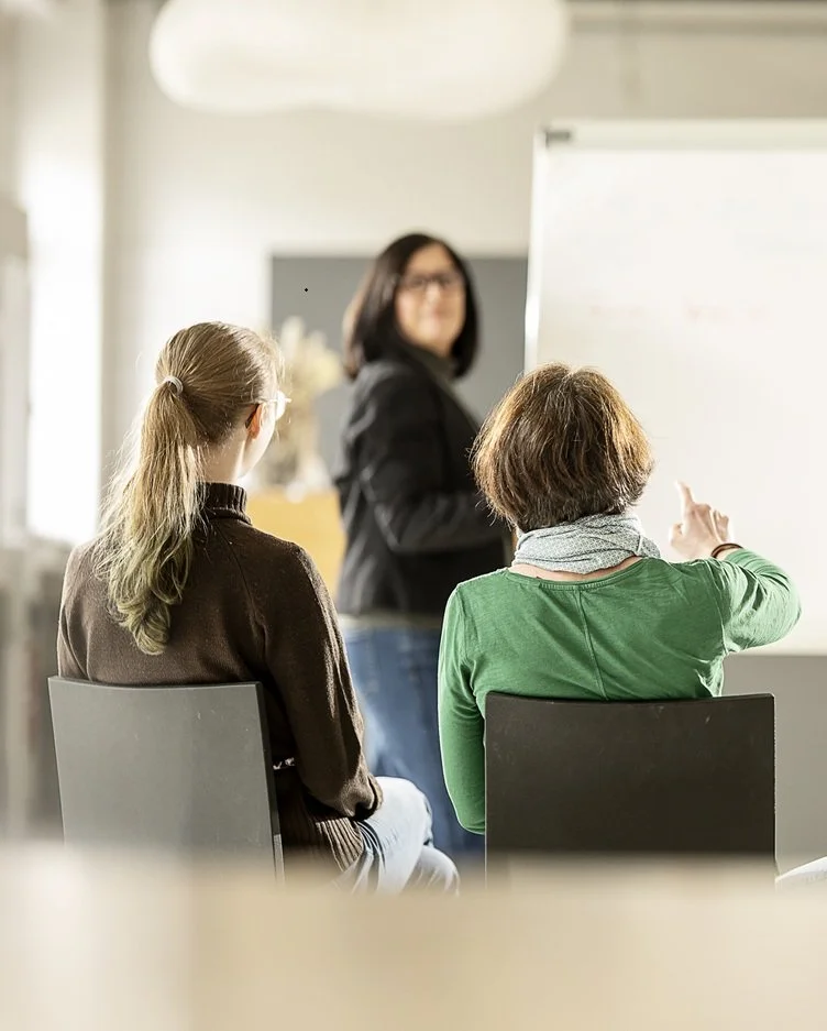 Zwei Frauen sitzen in einem Raum, während eine andere Frau am Whiteboard steht und erklärt.