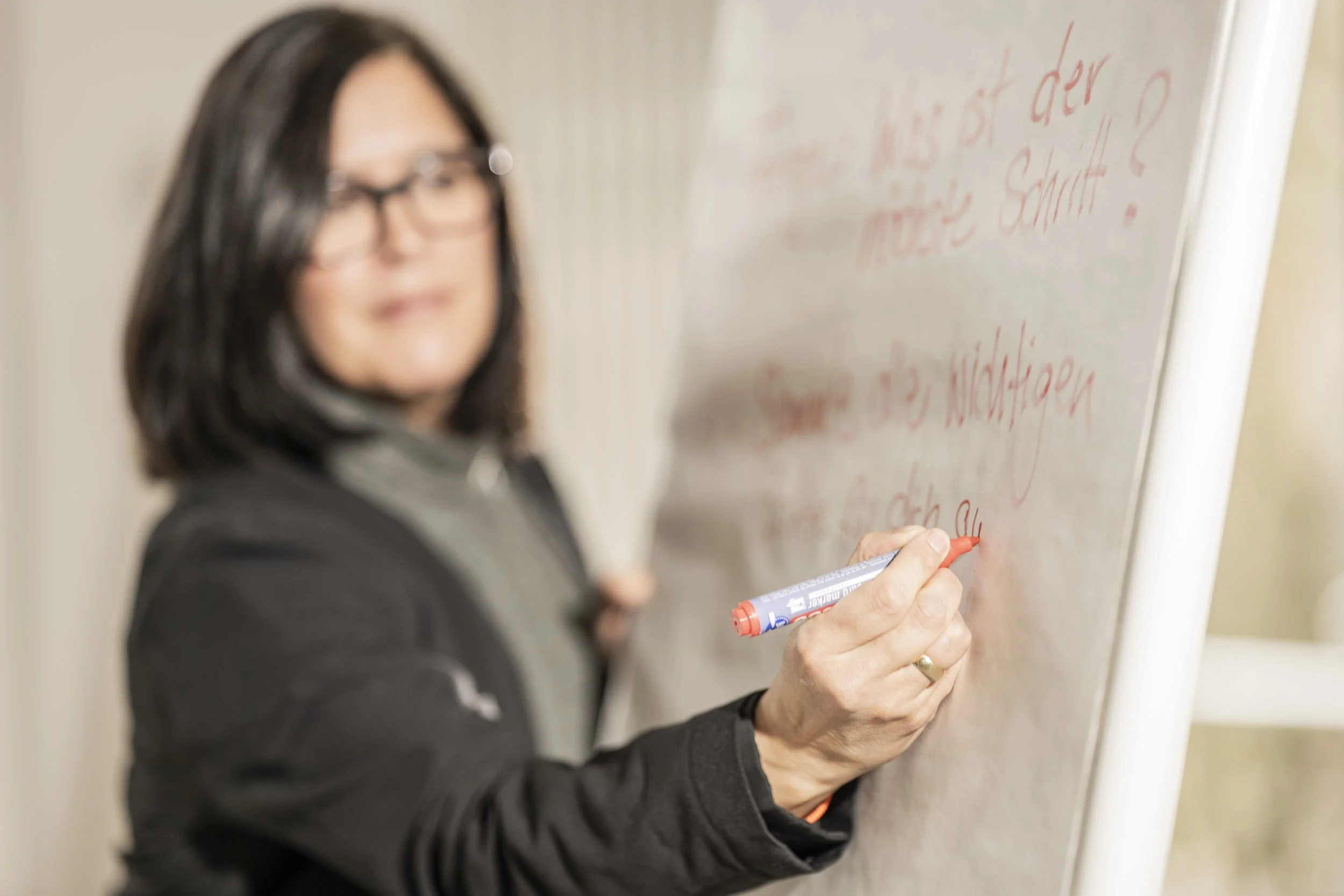 Eine Frau schreibt auf eine weiße Tafel mit einem roten Marker. Die Frau trägt eine schwarze Jacke und Brille. Der Text auf der Tafel ist unscharf und kaum lesbar.