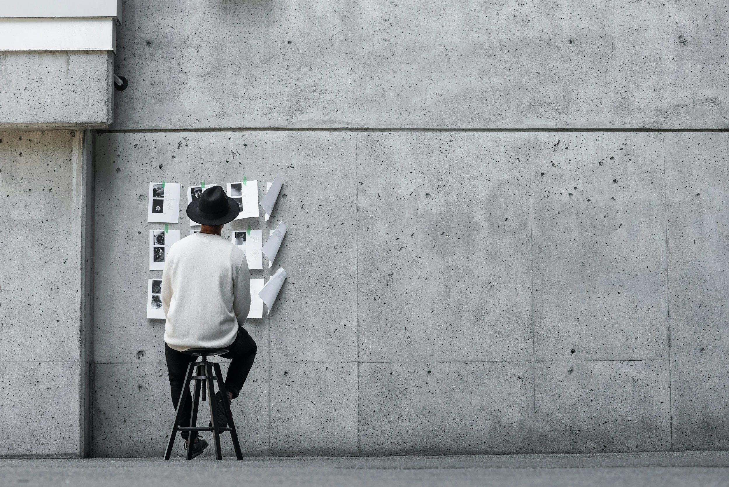 A person wearing a black hat and white shirt is sitting on a stool in front of a brick wall, looking at a display of printed photographs or documents taped to the wall.