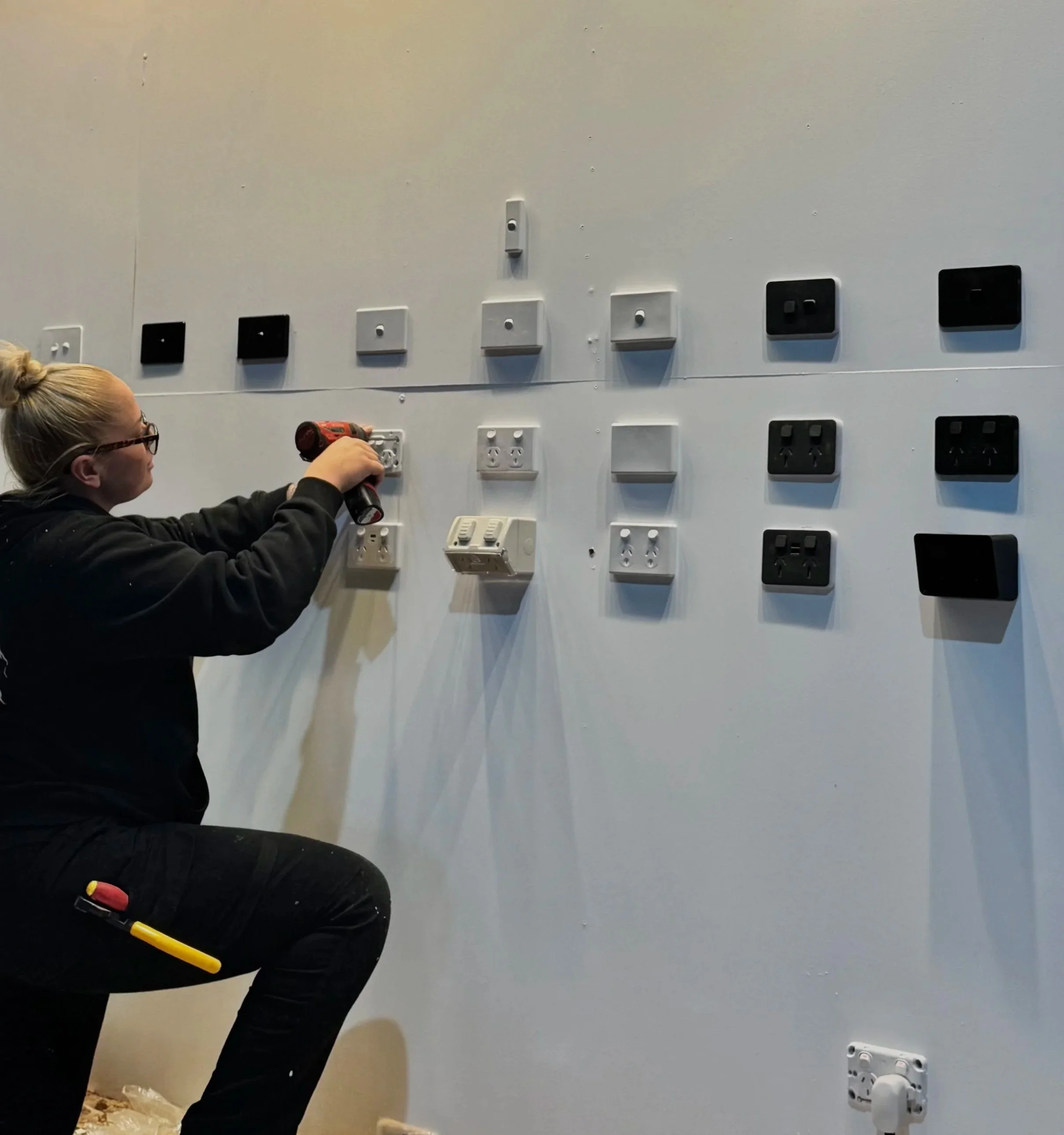 A person in black clothing is installing electrical outlets and switches on a white wall in a room that is under construction.