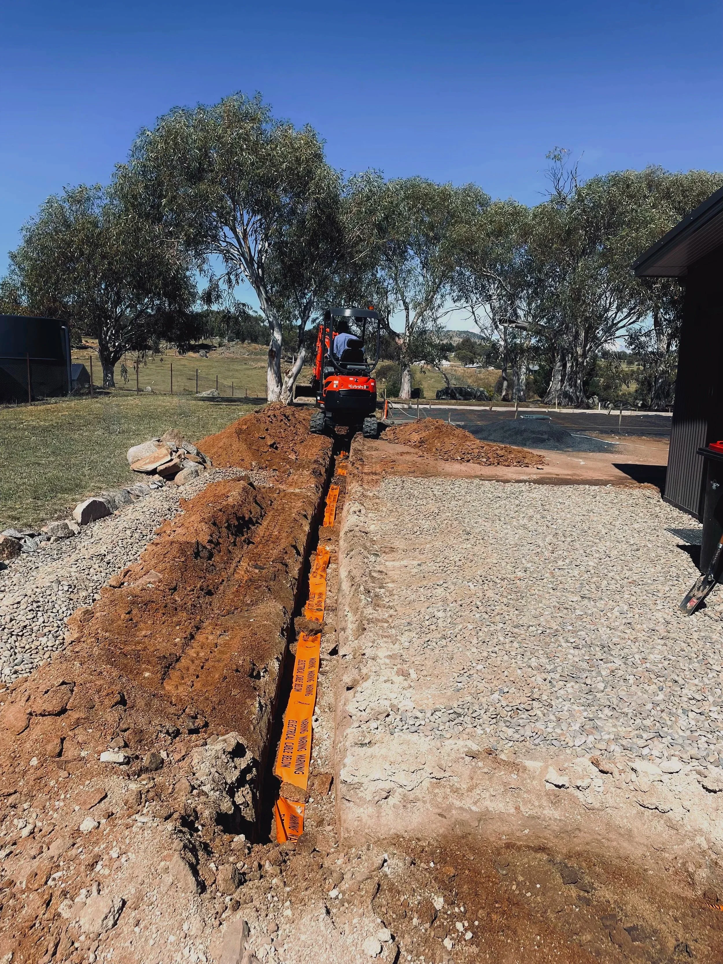 Construction site with a small excavator digging a trench, orange caution tape, and large trees in the background under a clear blue sky.