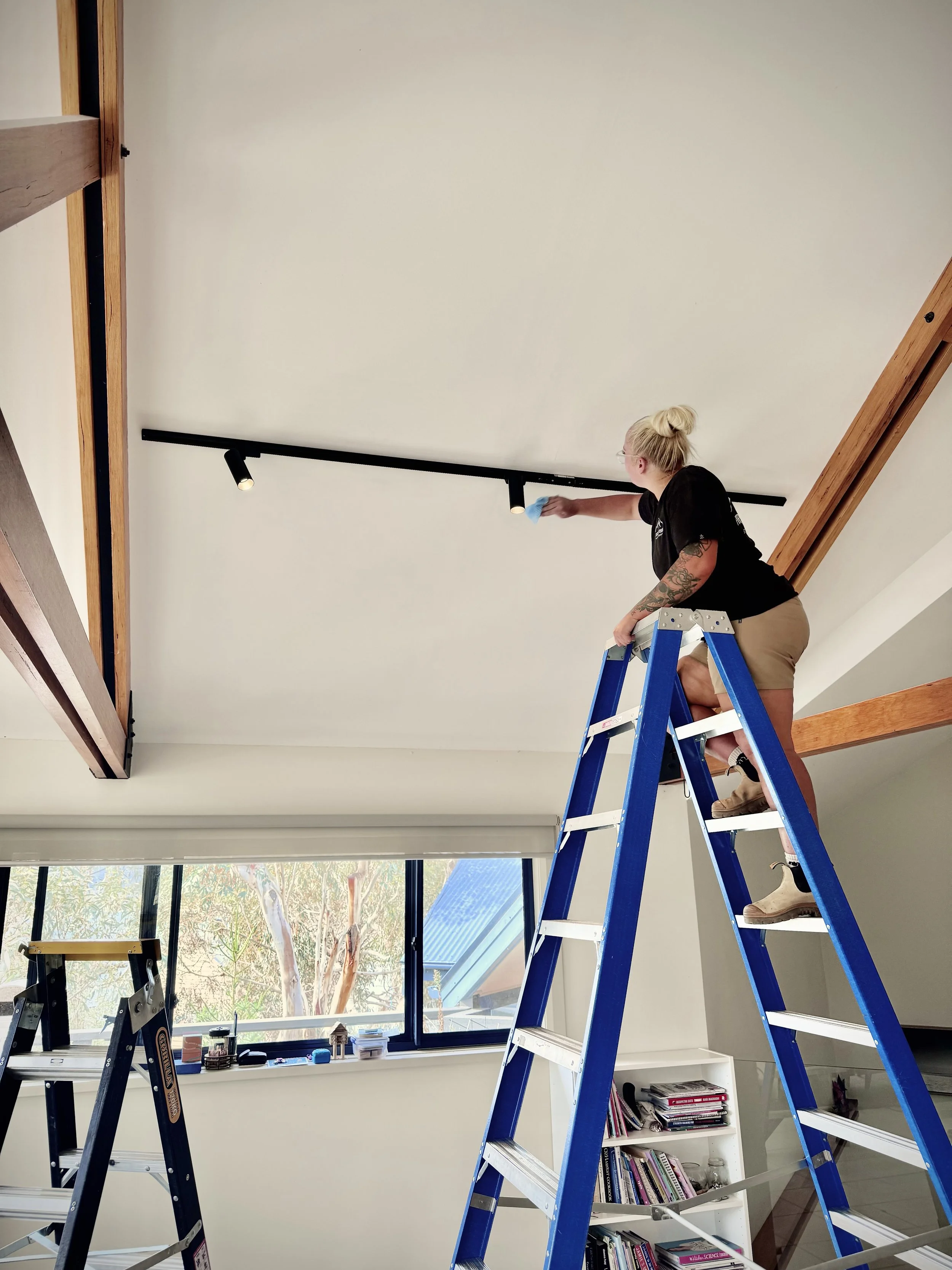A woman on a blue ladder cleaning a ceiling track light fixture in a modern room with large windows, wooden beams, and a bookshelf.