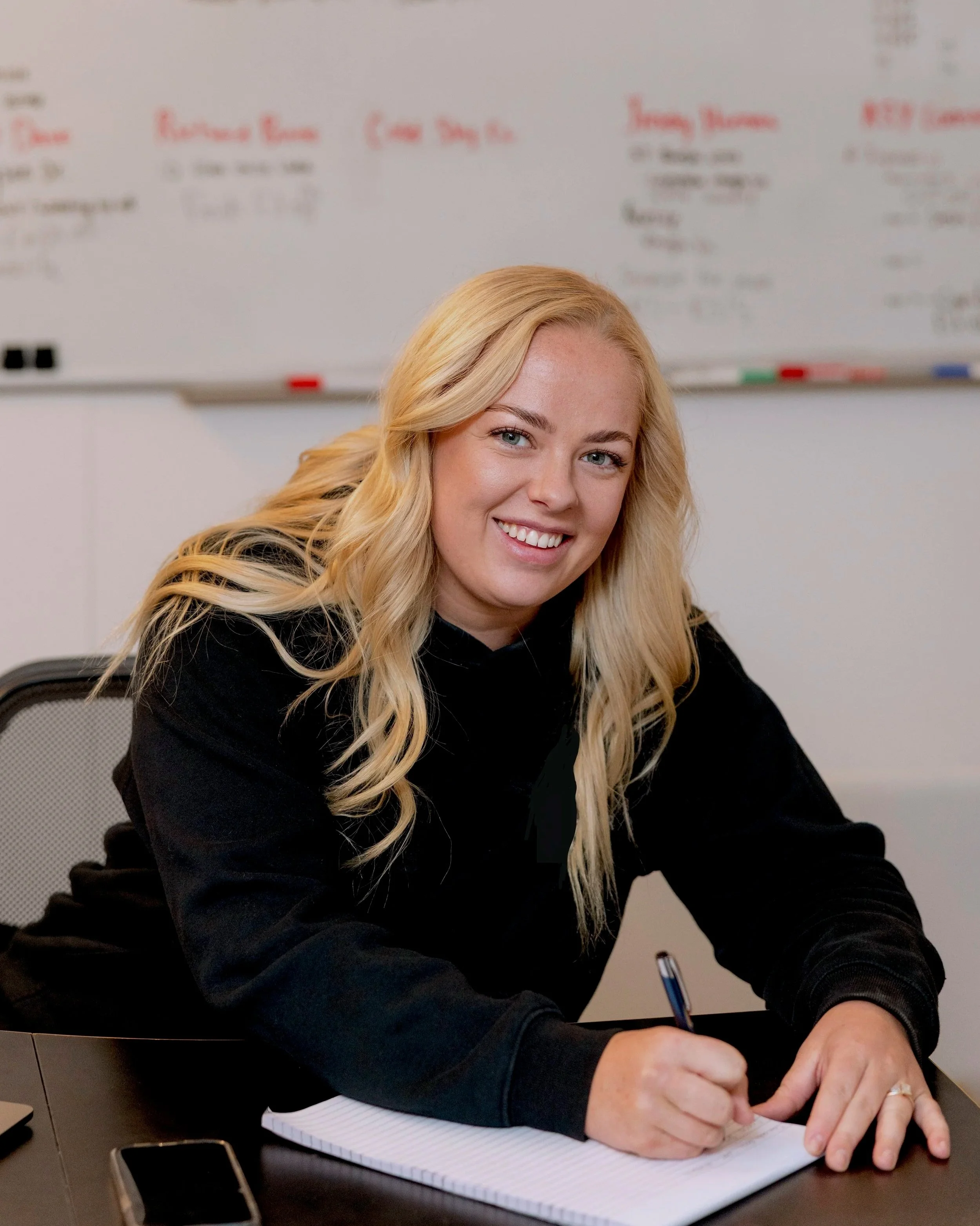 A woman with long blond hair, sitting at a desk, smiling, and writing in a notebook with a pen. Behind her is a large whiteboard with red and black writing.