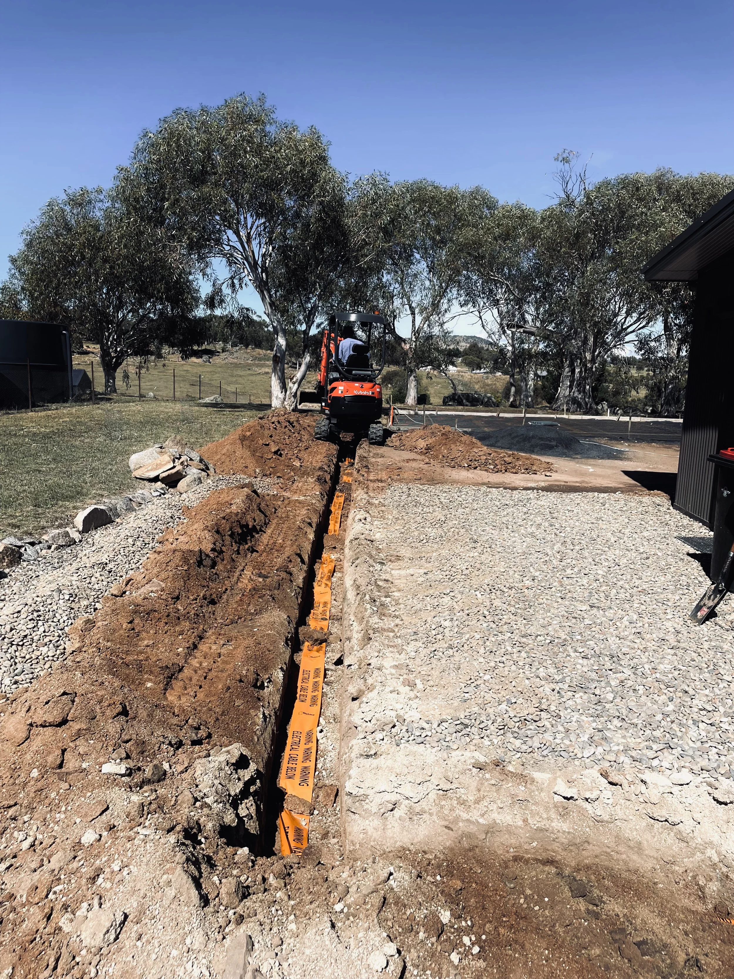 Construction site with an excavator digging a trench, orange warning tape along the trench, trees and open land in the background under a clear blue sky.
