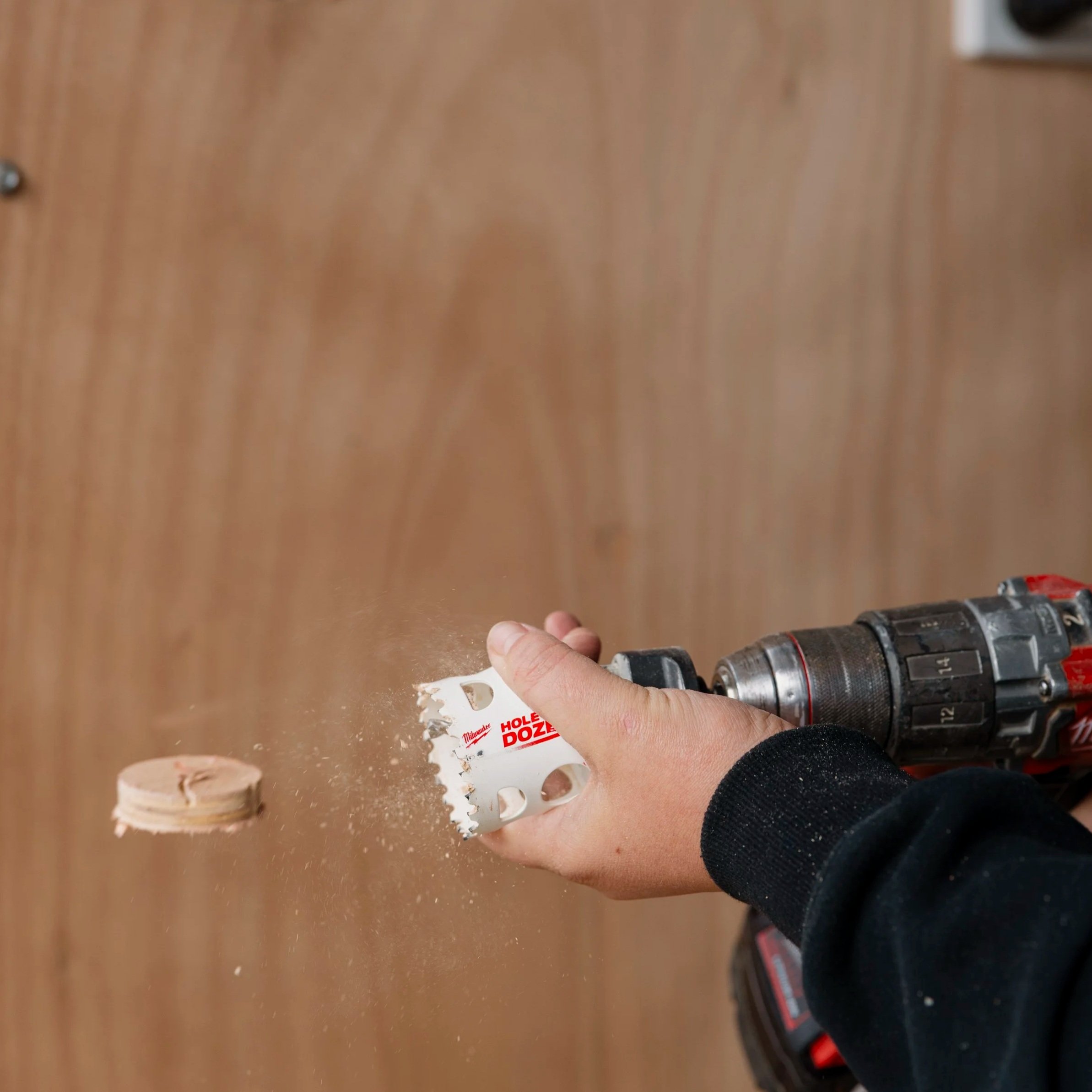A person using a cordless drill to secure a hole saw attachment into a piece of wood. The person's hand is visible, holding the drill, and a hole saw is cutting into the wood, with sawdust nearby. The background is a wood surface or workbench.