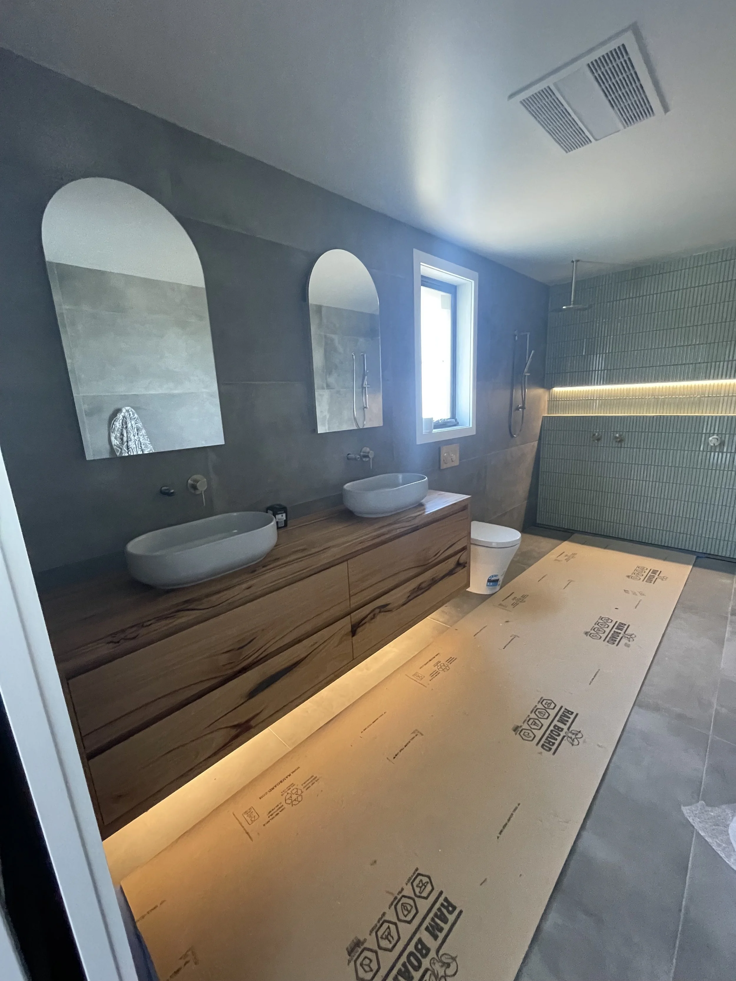 A modern bathroom under construction with two oval vessel sinks on a wooden vanity, mirrors above, a window, and a tiled shower area with a drain and a showerhead. Protective cardboard on the floor and an illuminated strip light.
