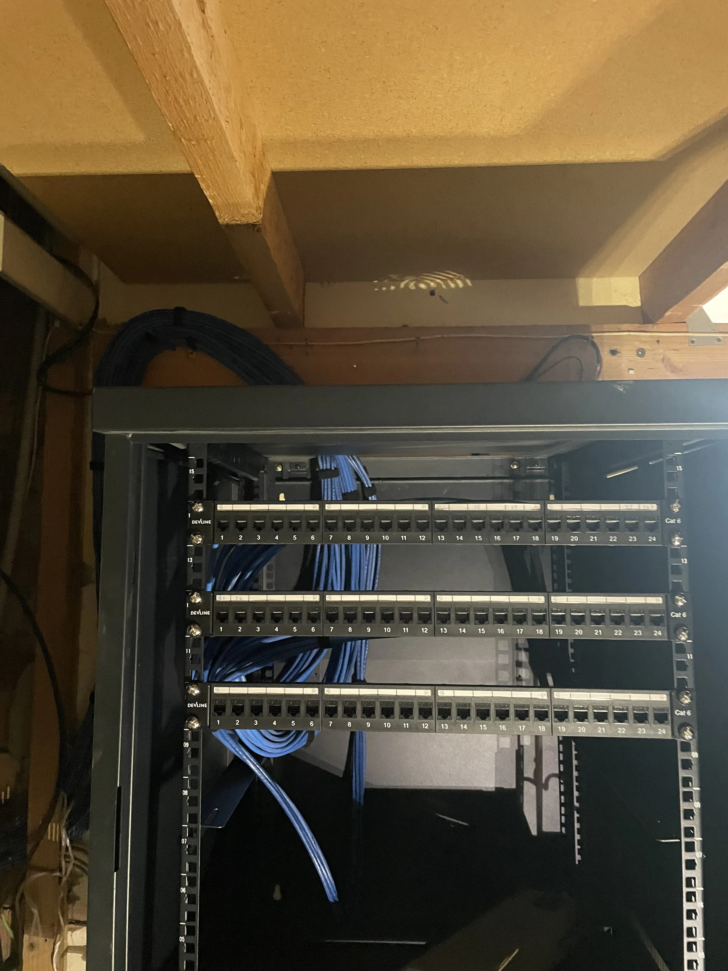 A server rack with a patch panel and blue Ethernet cables connected inside a metal frame, positioned on a black surface, with a wooden ceiling and wall in the background.