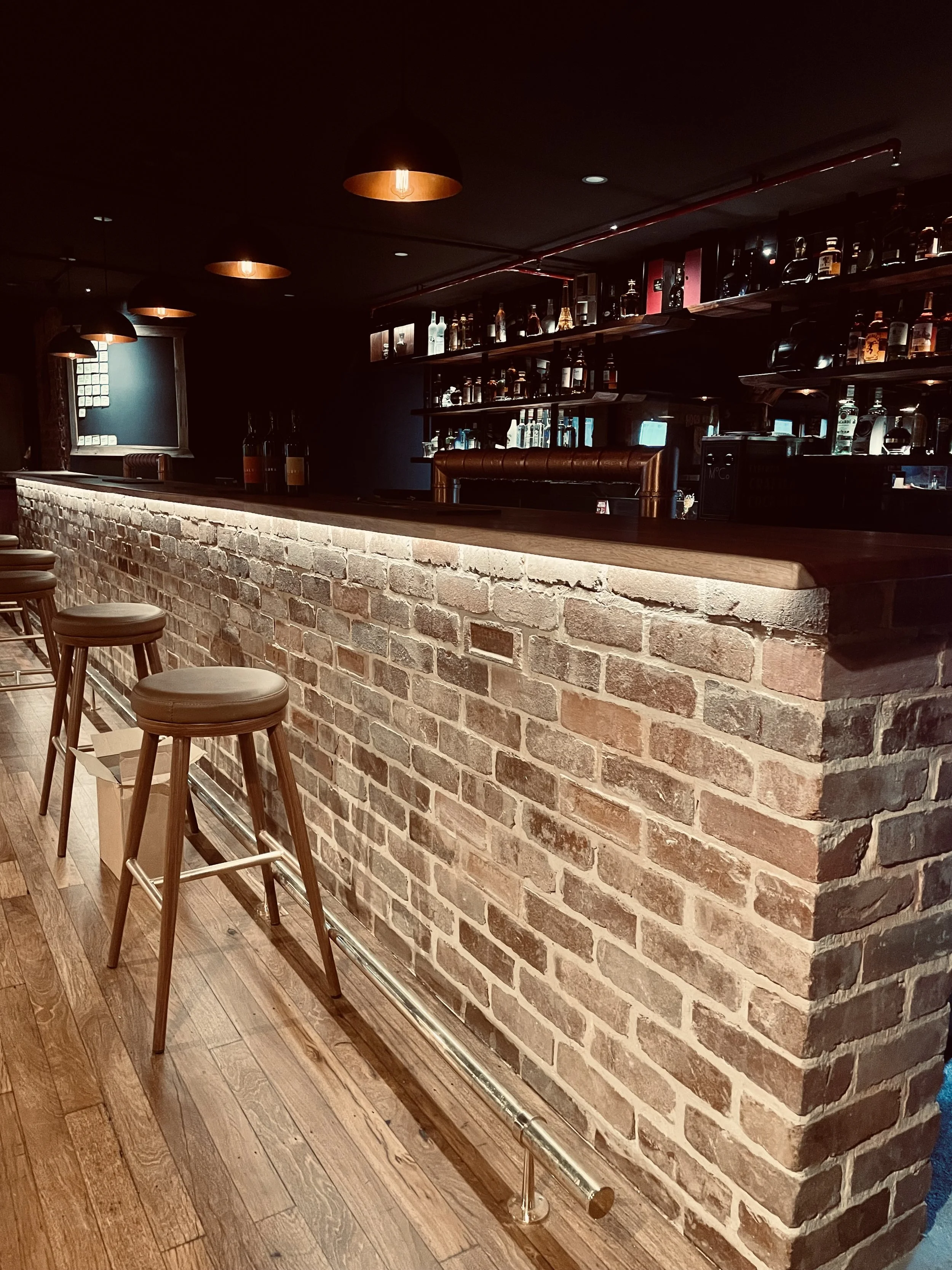 A dimly lit bar with a front brick counter and several bar stools. Behind the counter are shelves stocked with various bottles of alcohol, and overhead lights cast a warm glow.