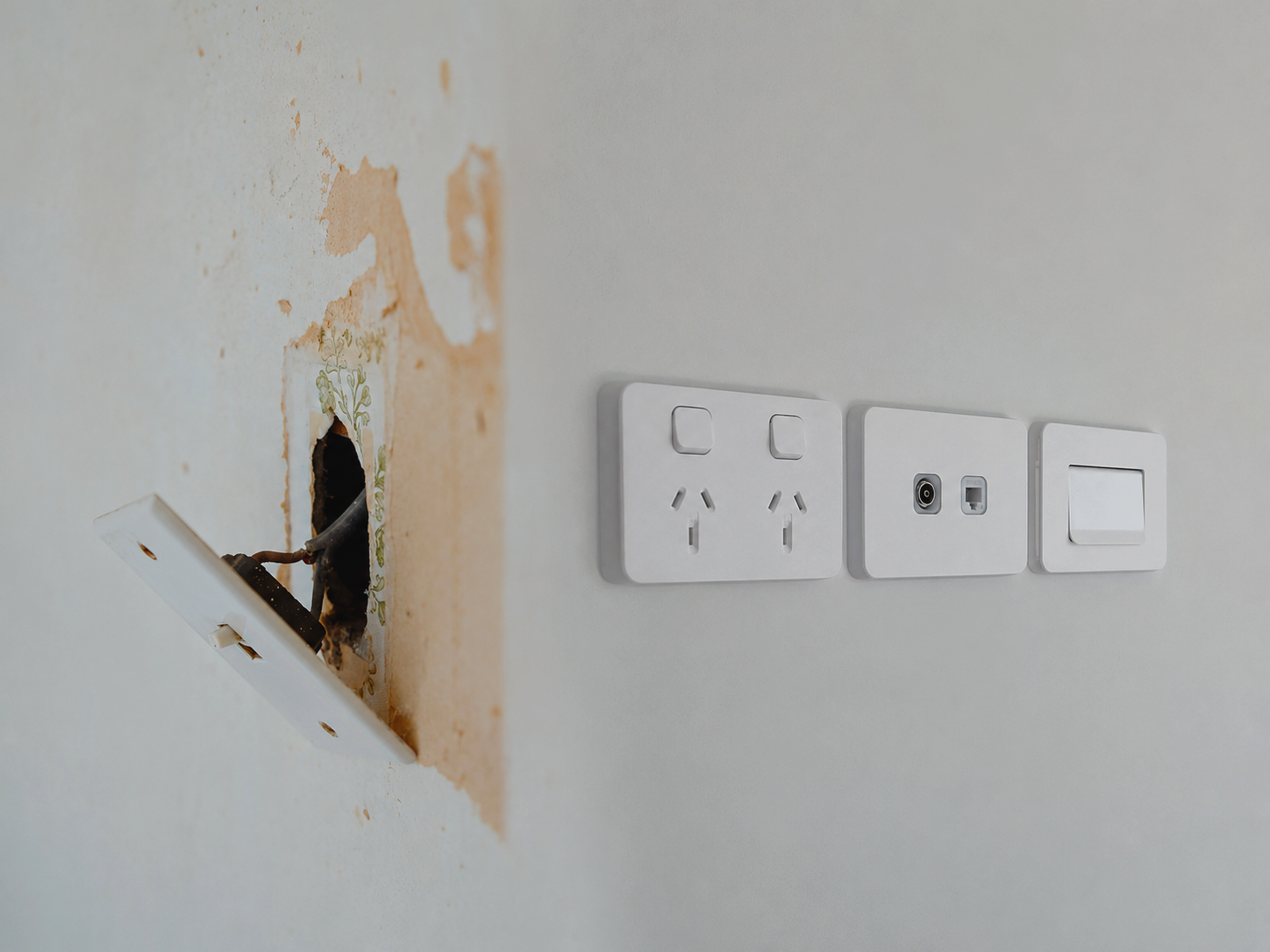 Close-up of a wall with a hole for a light switch, three new electrical outlets and switches installed next to it, with the surrounding wall partially stripped and exposed.