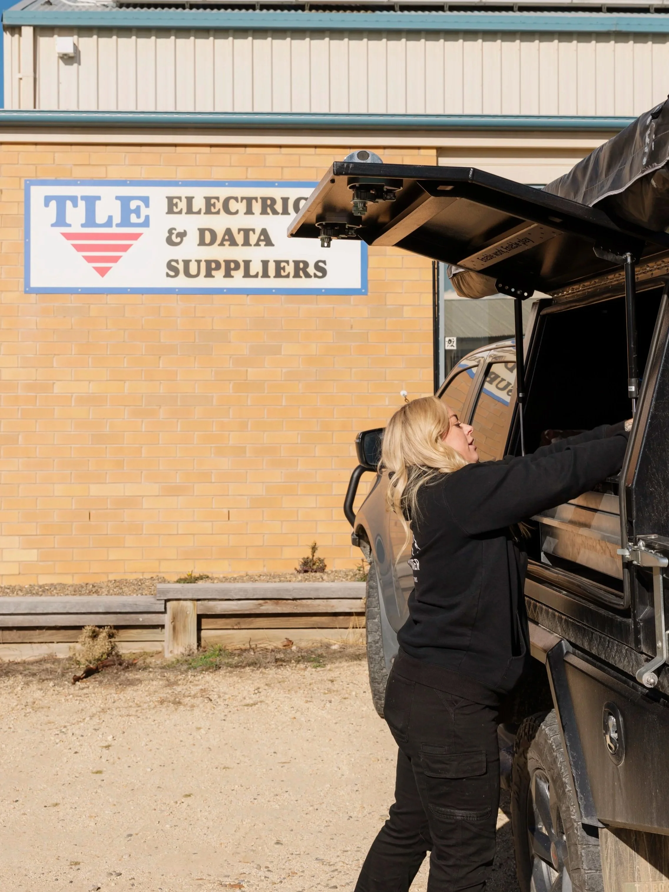 A woman with long blonde hair is standing next to a black off-road vehicle, leaning inside with her head and arms extended, seemingly working on or inspecting something inside the vehicle. The vehicle has an open roof and appears to be in a parking lot outside a building with a sign that reads 'TLE Electric & Data Suppliers.'