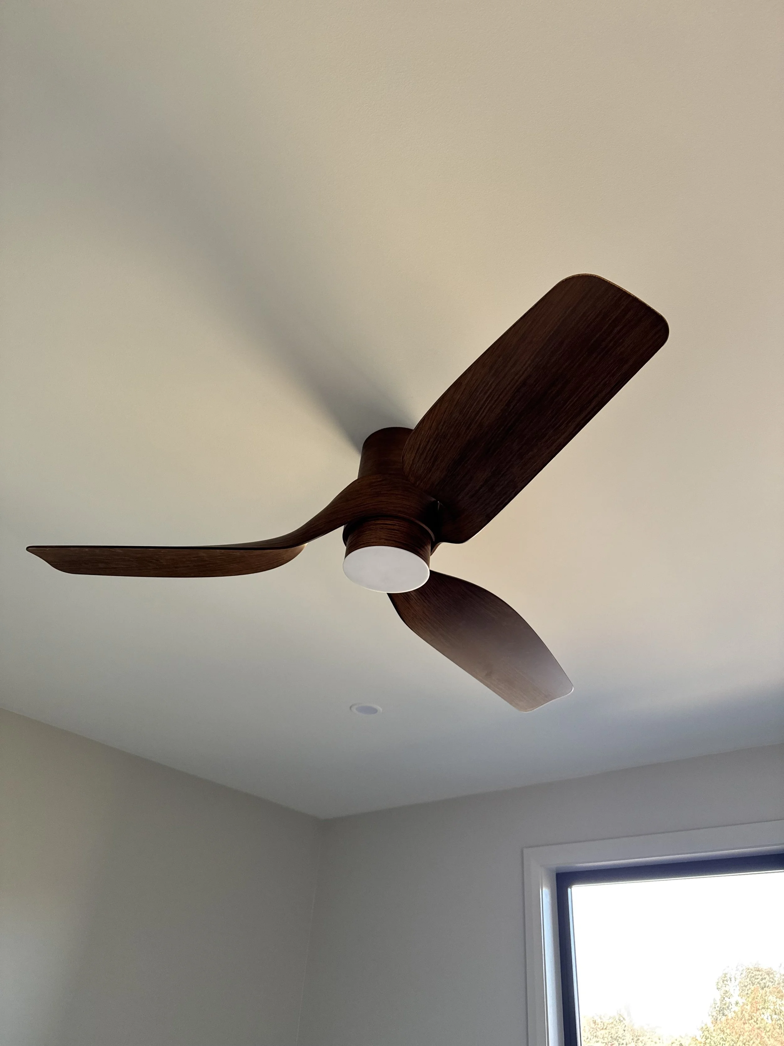 A ceiling fan with three wooden blades and a white light fixture in a room with a window showing trees outside.