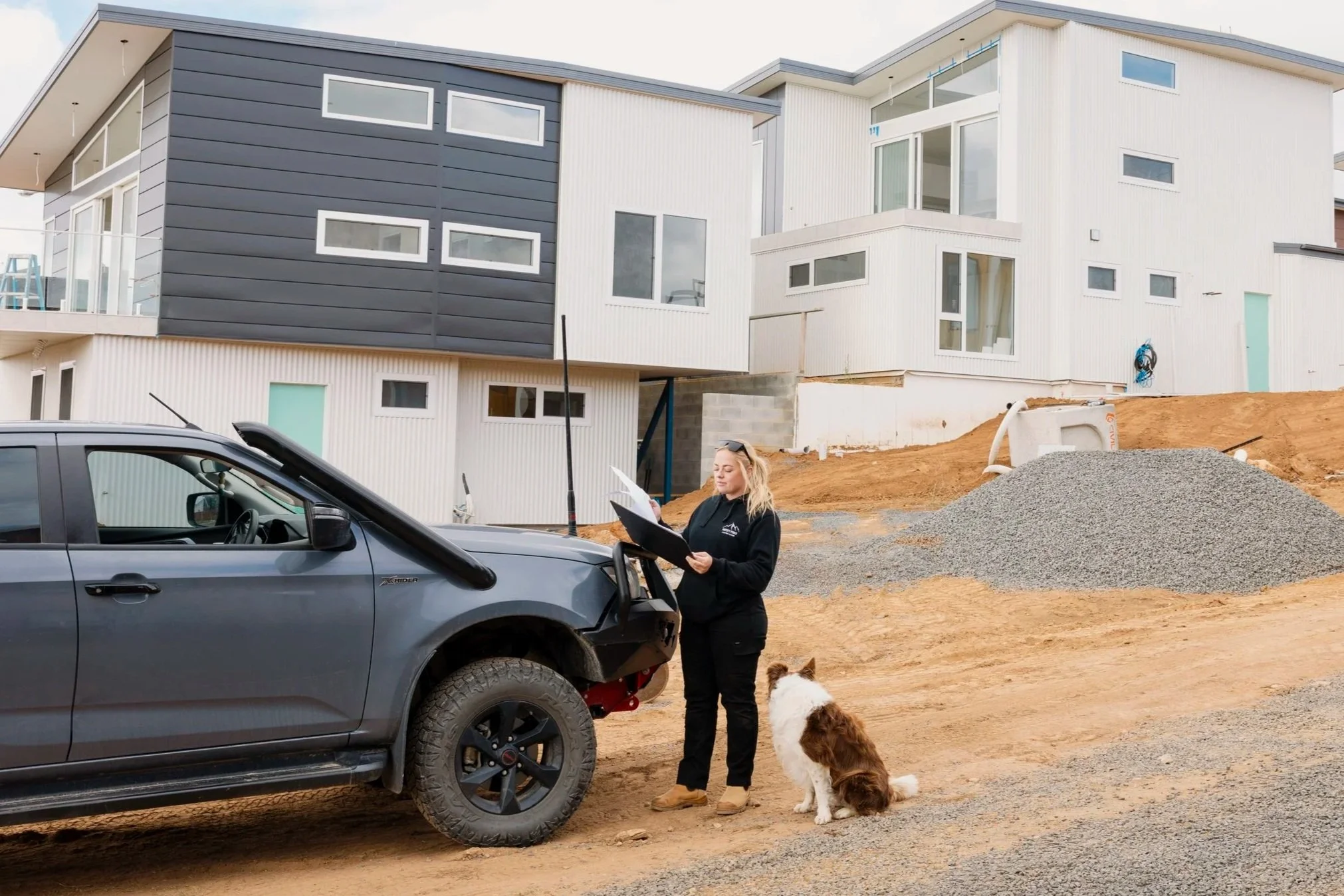 A woman standing next to a black pickup truck at a construction site, holding papers, with two dogs nearby, with modern residential buildings under construction in the background.