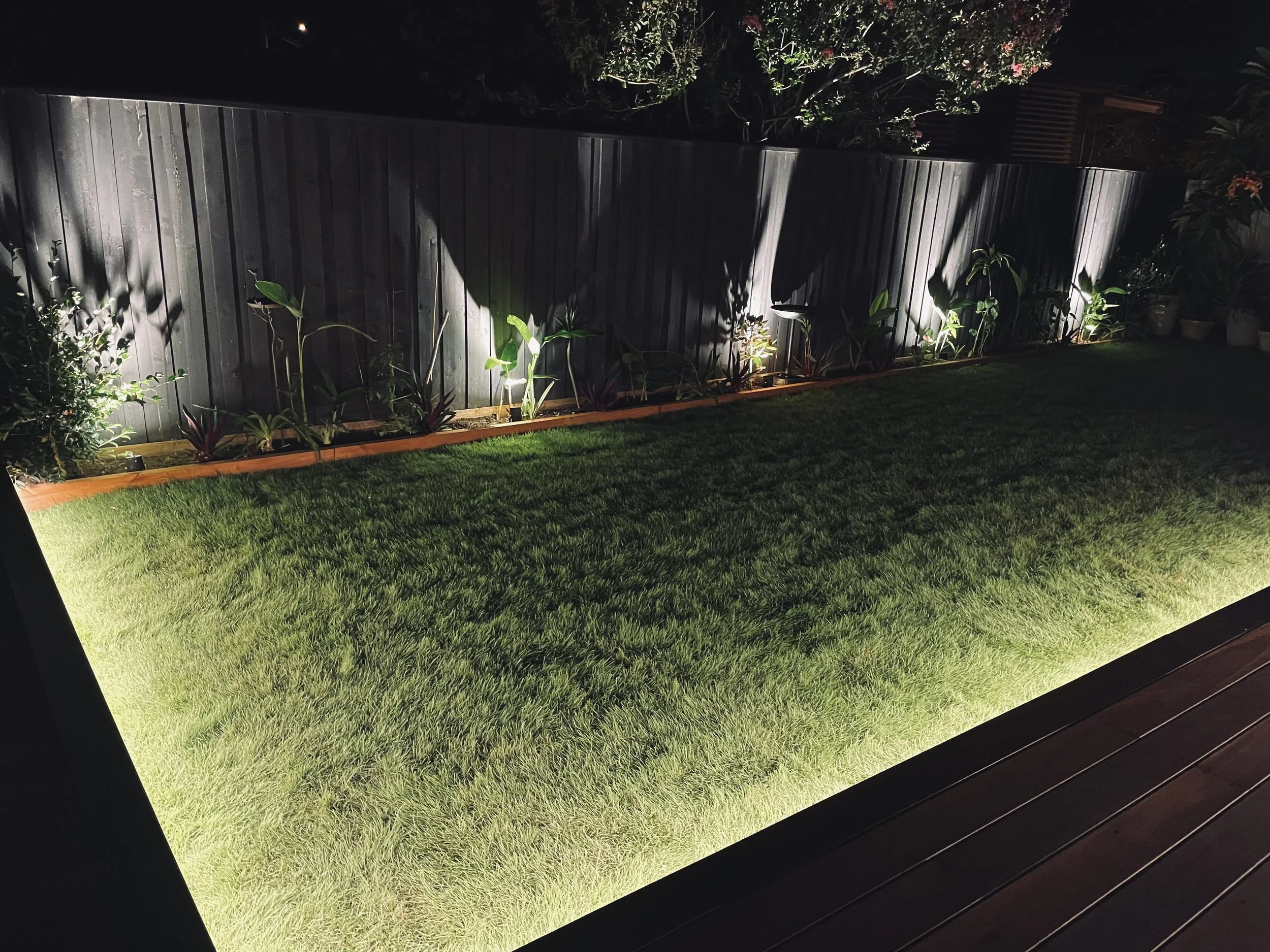 A backyard at night with a wooden fence, garden plants, and LED strip illuminated the grass/lawn.
