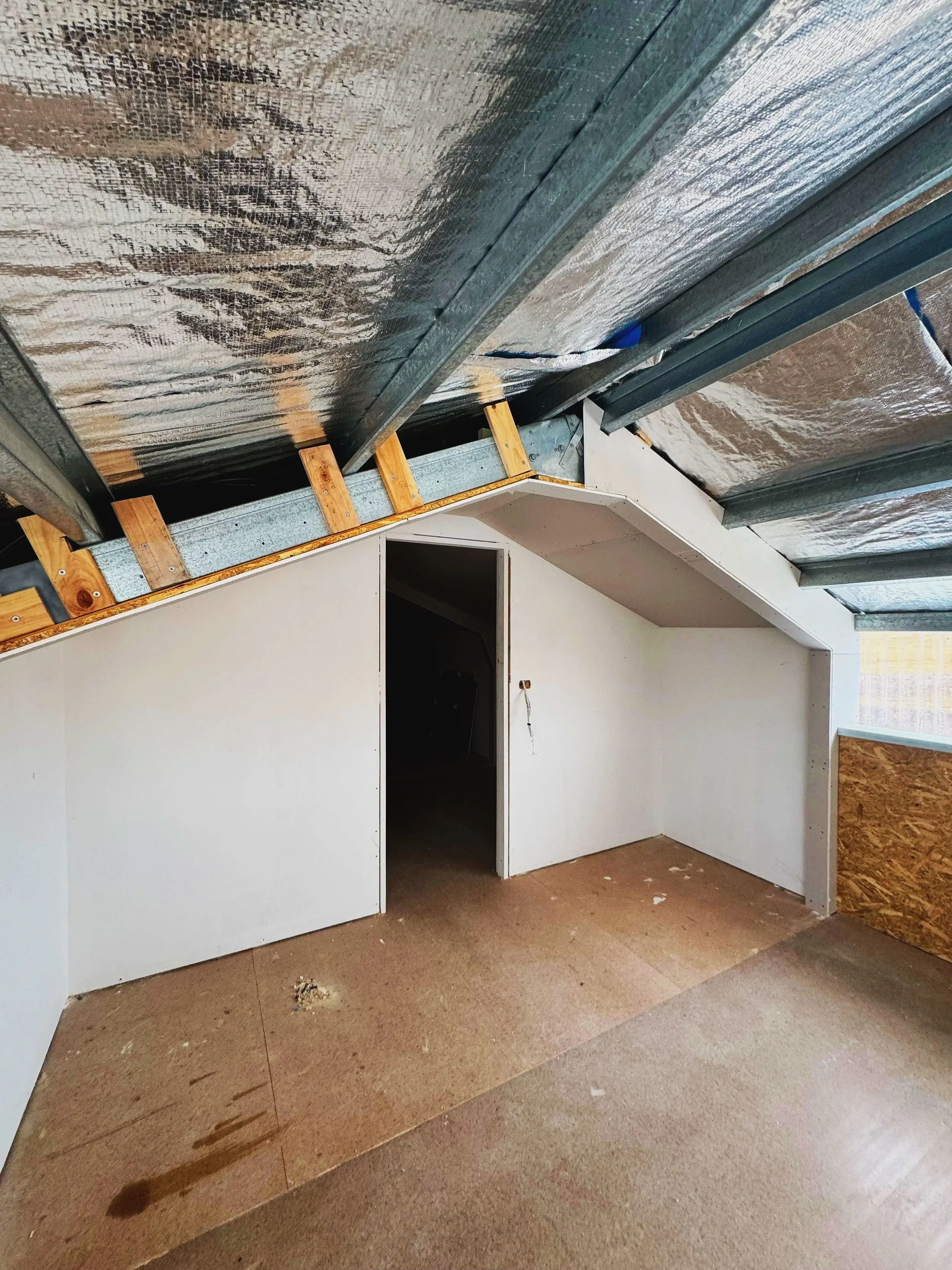 Interior under construction or renovation with drywall partially installed, exposed ceiling insulation, and unfinished flooring.