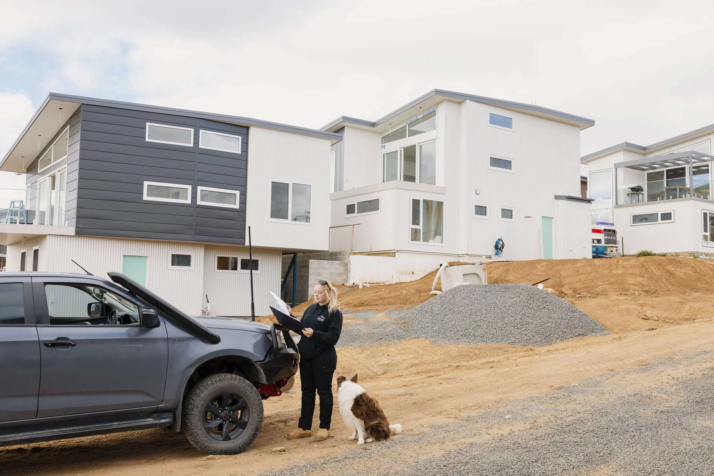 A woman with a dog standing next to a gray pickup truck on a construction site in front of modern white and gray buildings. The woman appears to be reviewing documents.